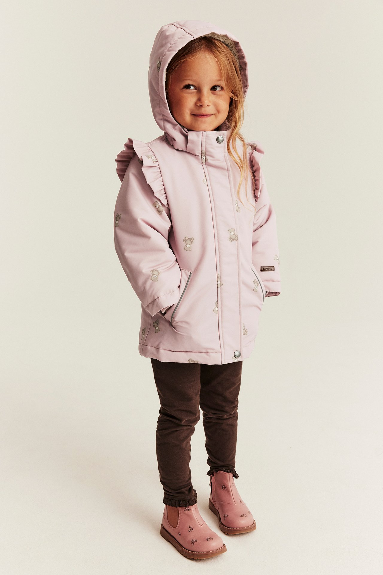 Front view of a kid wearing a pink hooded jacket with teddy bears, brown leggings, and pink boots.