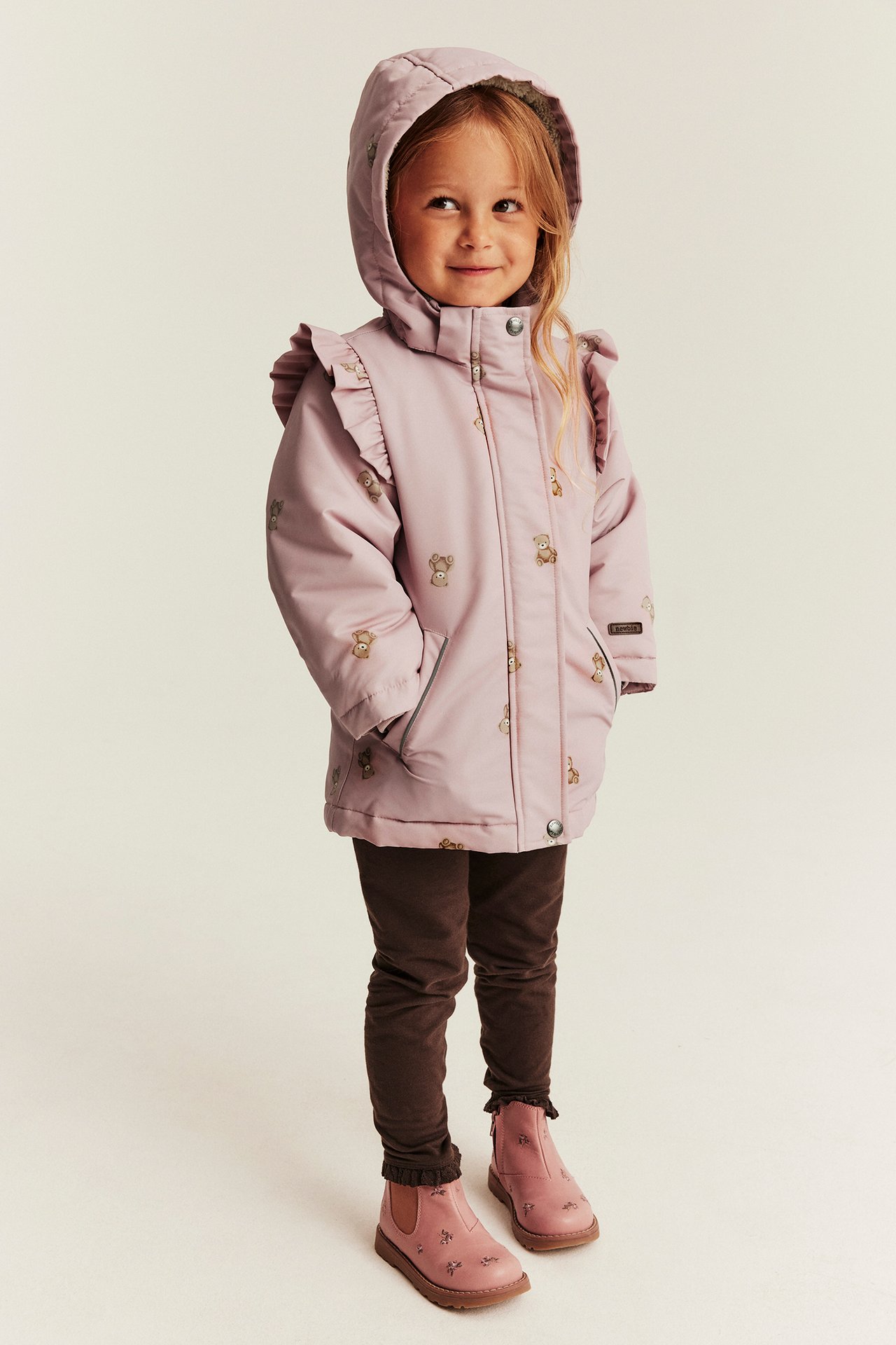 Front view: kid wears light pink hooded teddy-print jacket with ruffles, brown leggings, pink boots.