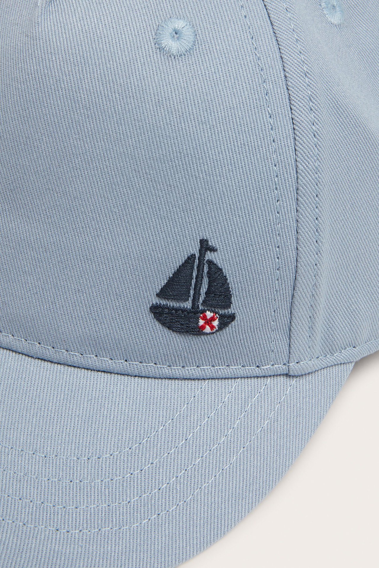 Close-up front view of a light blue baby/kid's cap with a dark blue embroidered sailboat motif.