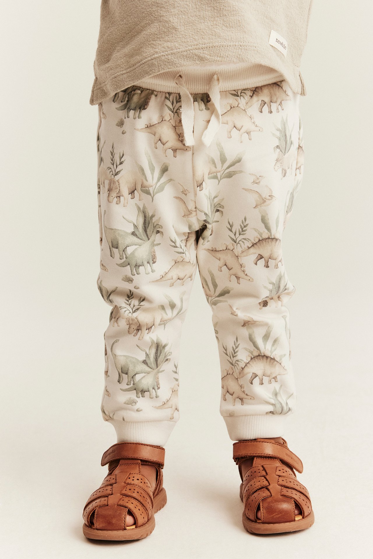 Vorderansicht von Baby in beiger Dinosaurier-Jogginghose, braunen Ledersandalen und beigem Langarmshirt.