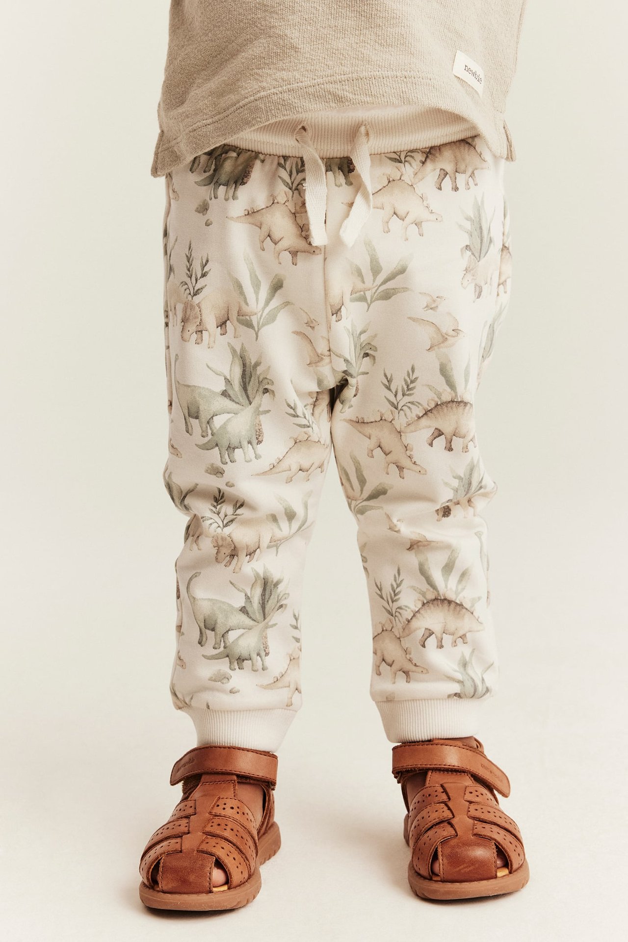 Vorderansicht von Baby in beiger Dinosaurier-Jogginghose, braunen Ledersandalen und beigem Langarmshirt.