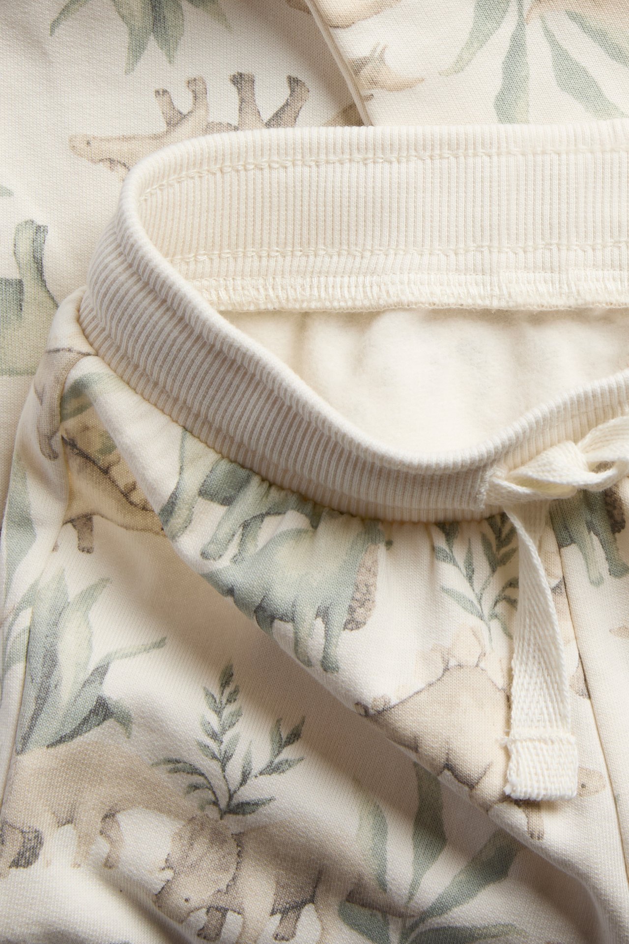 Nahaufnahme von hellbeiger Kinderhose mit Dino-Muster, geripptem Bund und Kordelzug.