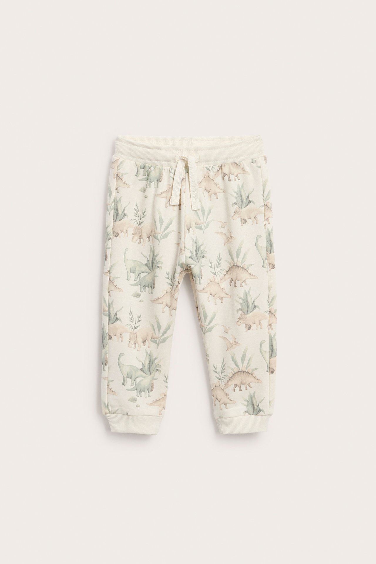 Vorderansicht: Baby-Jogginghose in Creme mit Dinosaurier- und Pflanzenmuster, Kordelzug und Bündchen.