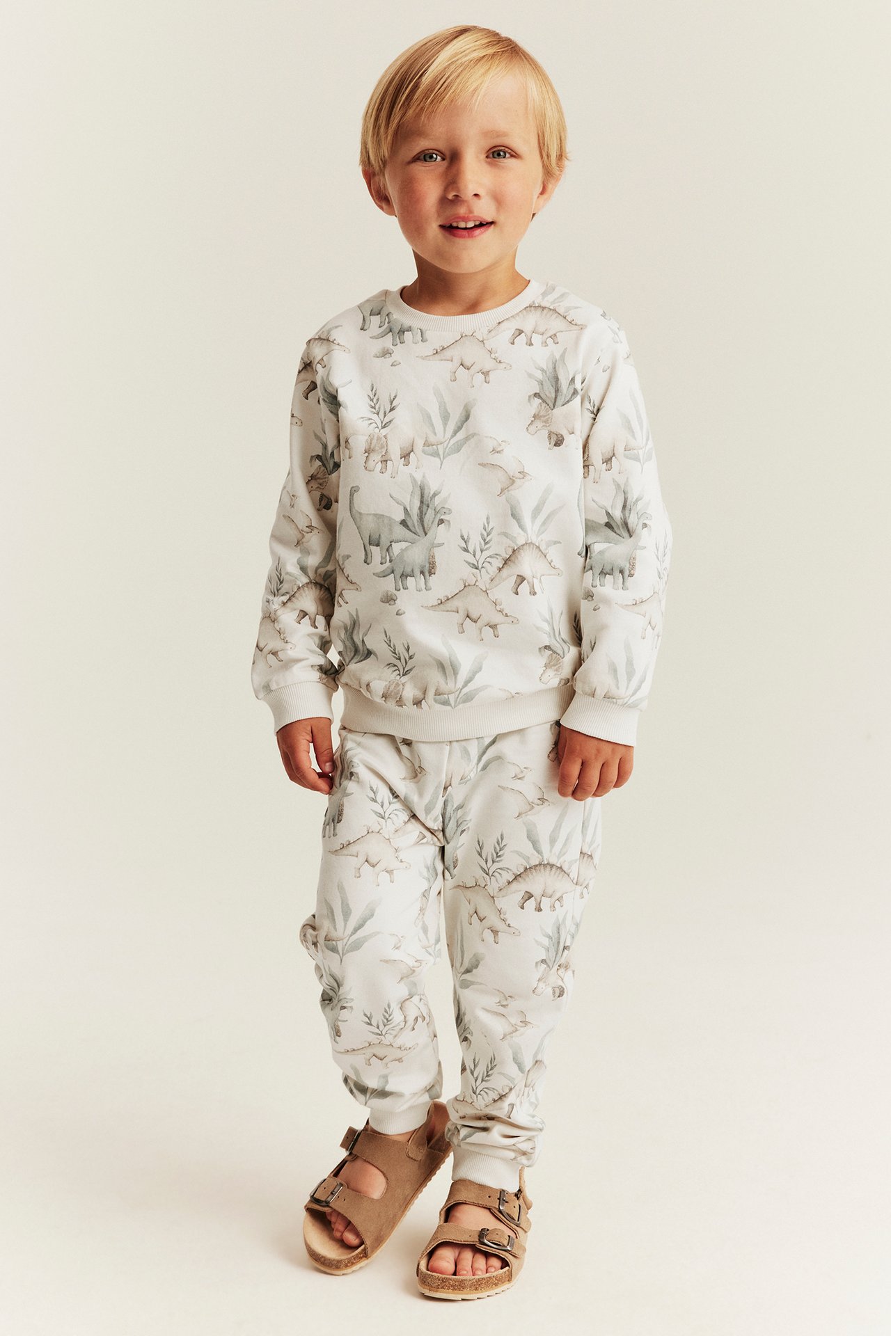 Frontansicht eines Jungen in weißem Dino-Print-Sweatshirt und Jogginghose mit braunen Sandalen.