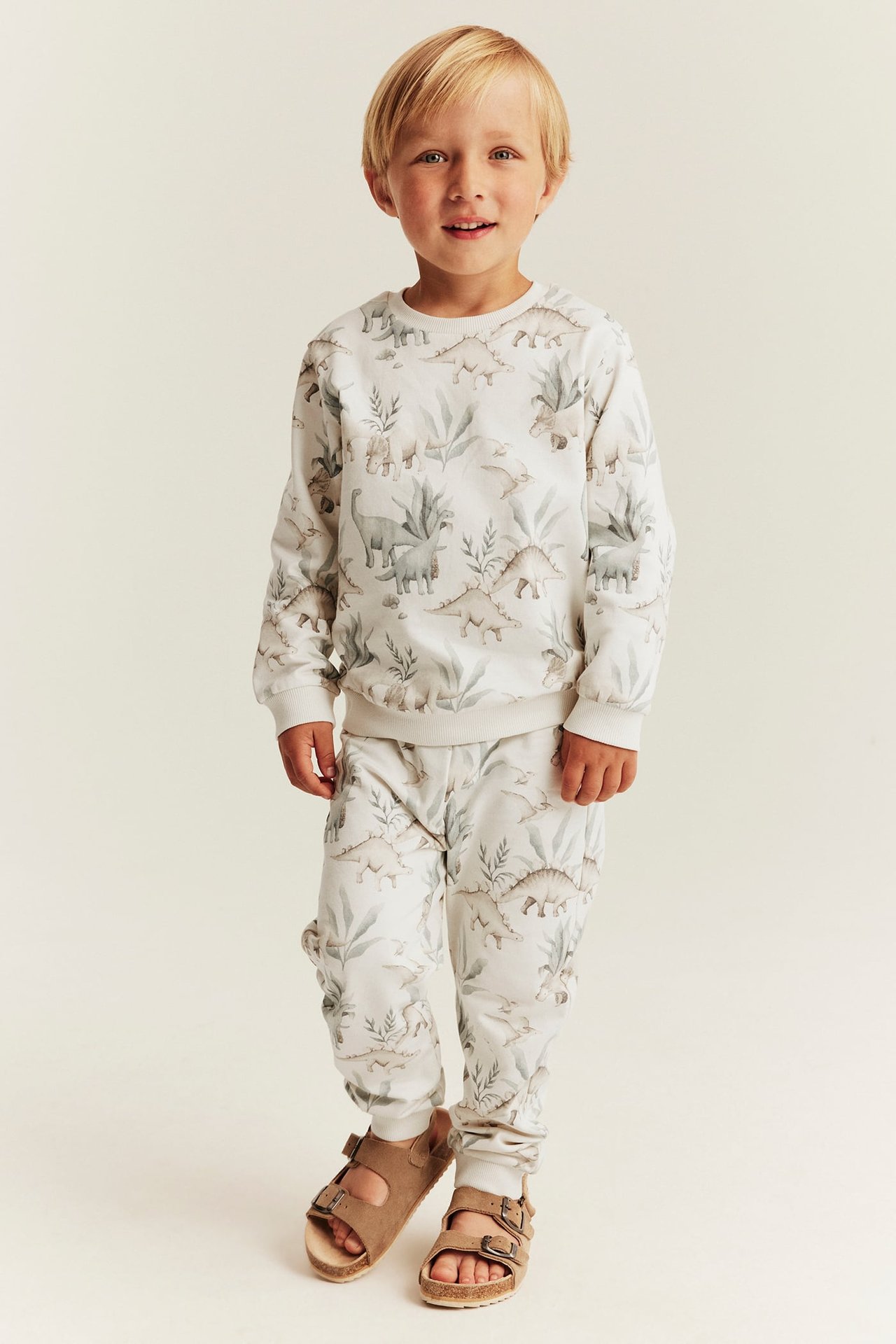 Frontansicht eines Jungen in weißem Dino-Print-Sweatshirt und Jogginghose mit braunen Sandalen.