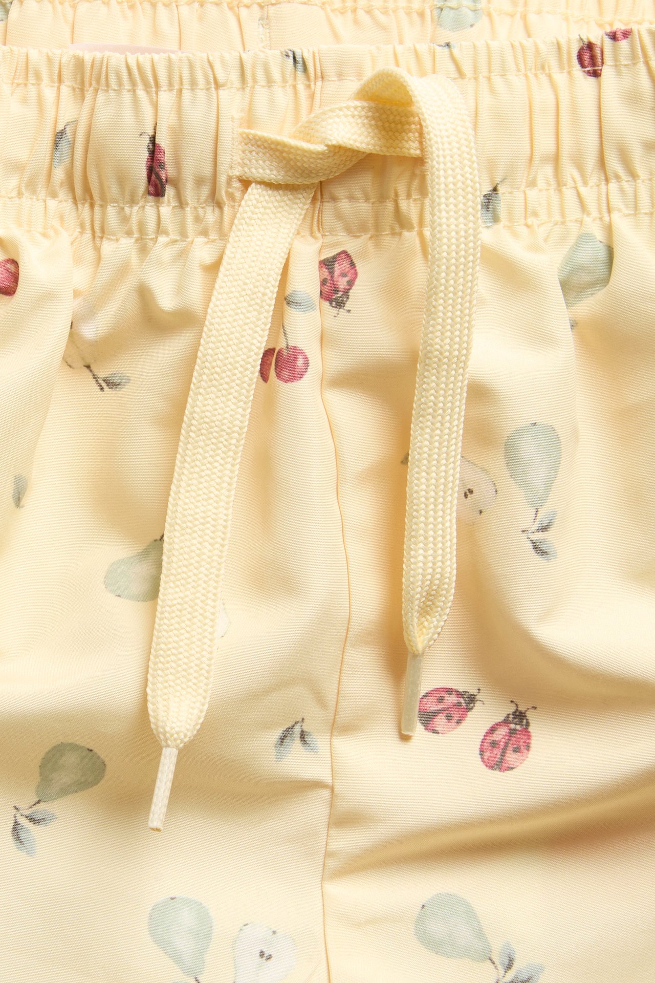 Close-up of kid's yellow swim shorts with ladybug, cherry, pear pattern and a drawstring waistband.