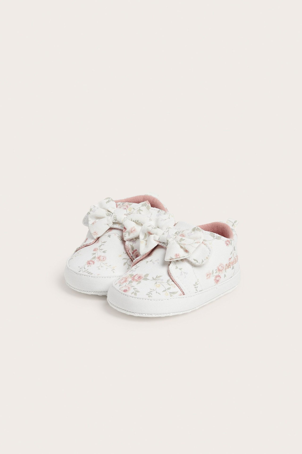 Vorderansicht von off-white Babyschuhen mit Blumenmuster, Klettverschluss, Stoffschleifen und rosa Futter.