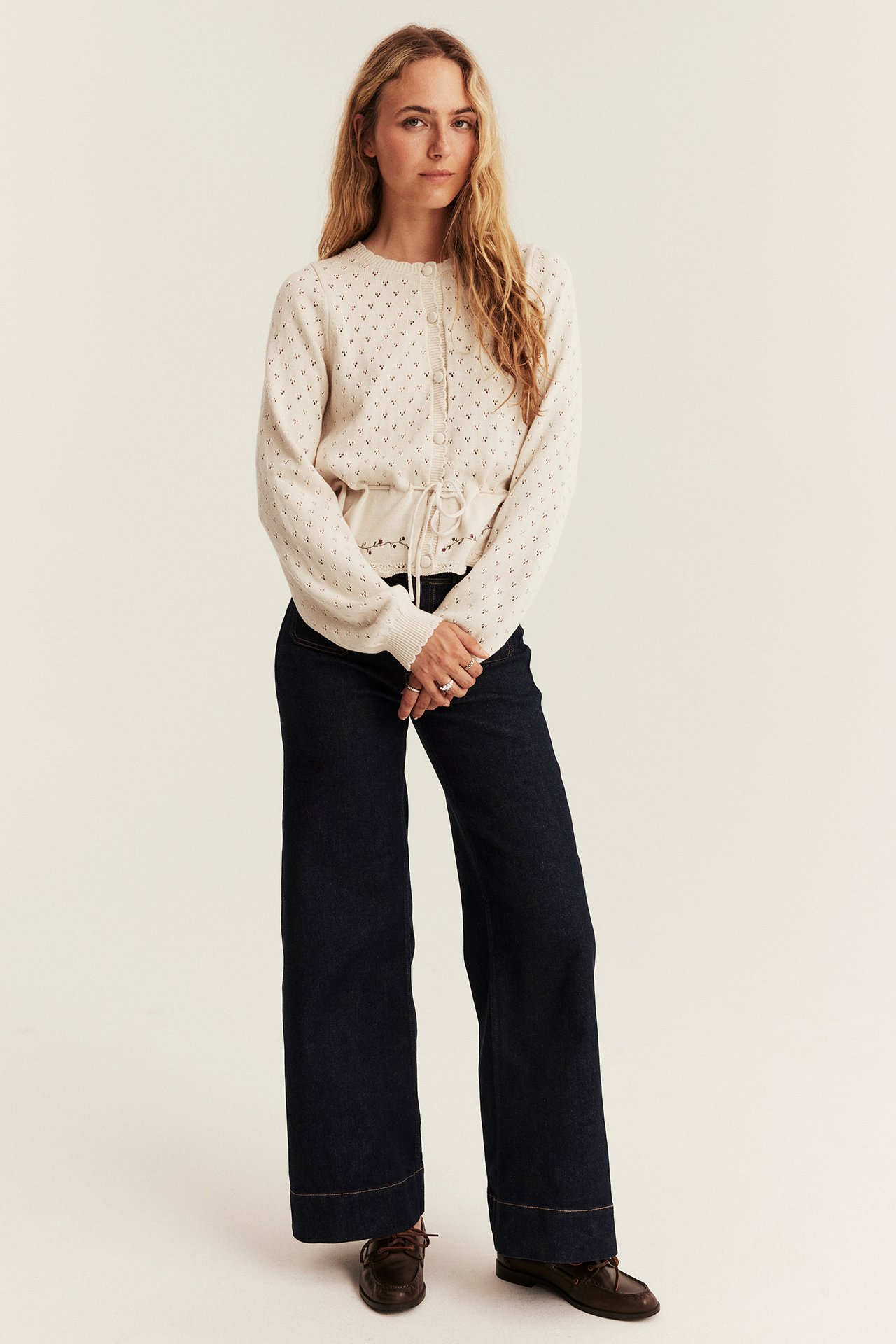 Vorderansicht, weibliches Modell in cremefarbener Lochstrick-Cardigan, dunkler Jeans und braunen Loafern.