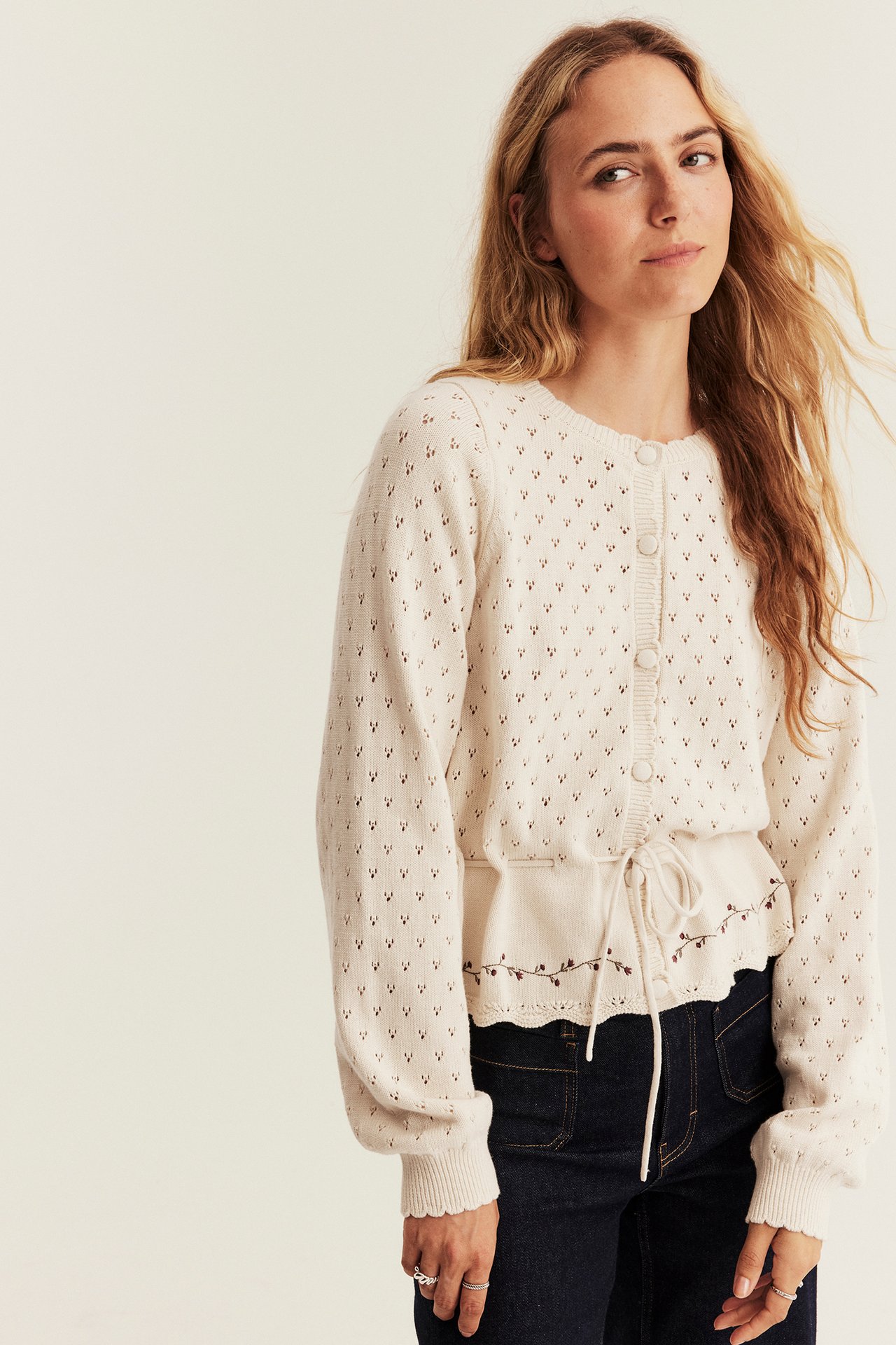 Vorderansicht einer Frau in cremefarbener Lochstrickjacke mit Bindeband, dunklen Jeans und Ringen.