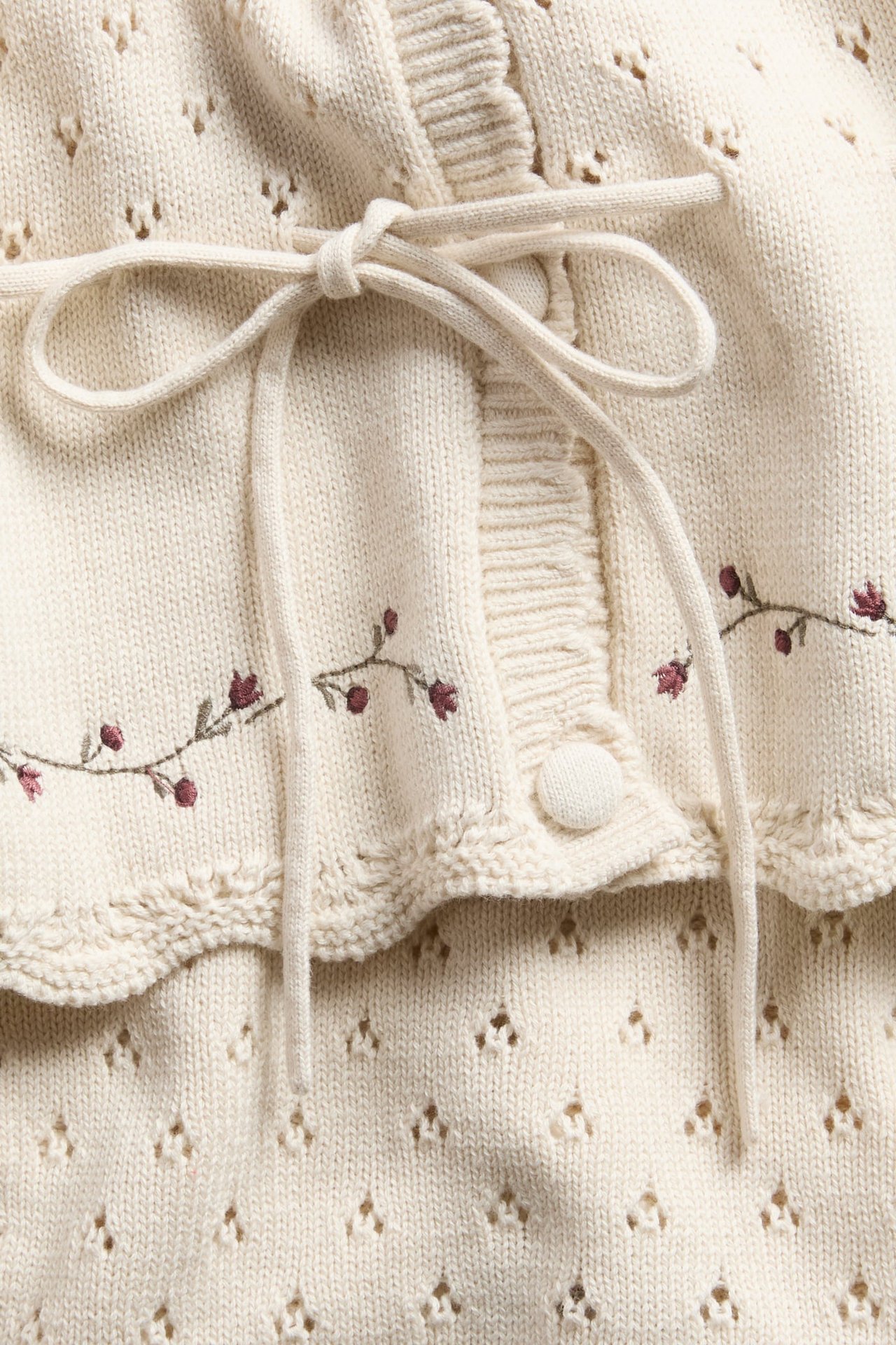 Nahaufnahme einer cremefarbenen Strickjacke mit Blumenstickerei und Schleife über Lochstrick-Kleidungsstück.