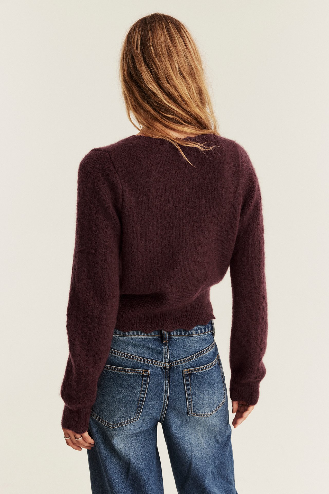 Rückenansicht einer Frau in dunkelrotem, flauschigem Strick-Cropped-Pullover mit Wellensaum, blauen Jeans.