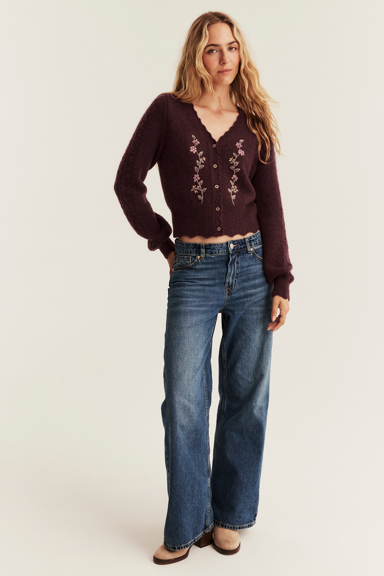 Vorderansicht: Frau in weinroter floral bestickter Strickjacke, blauer weiter Jeans und beigen Stiefeletten.