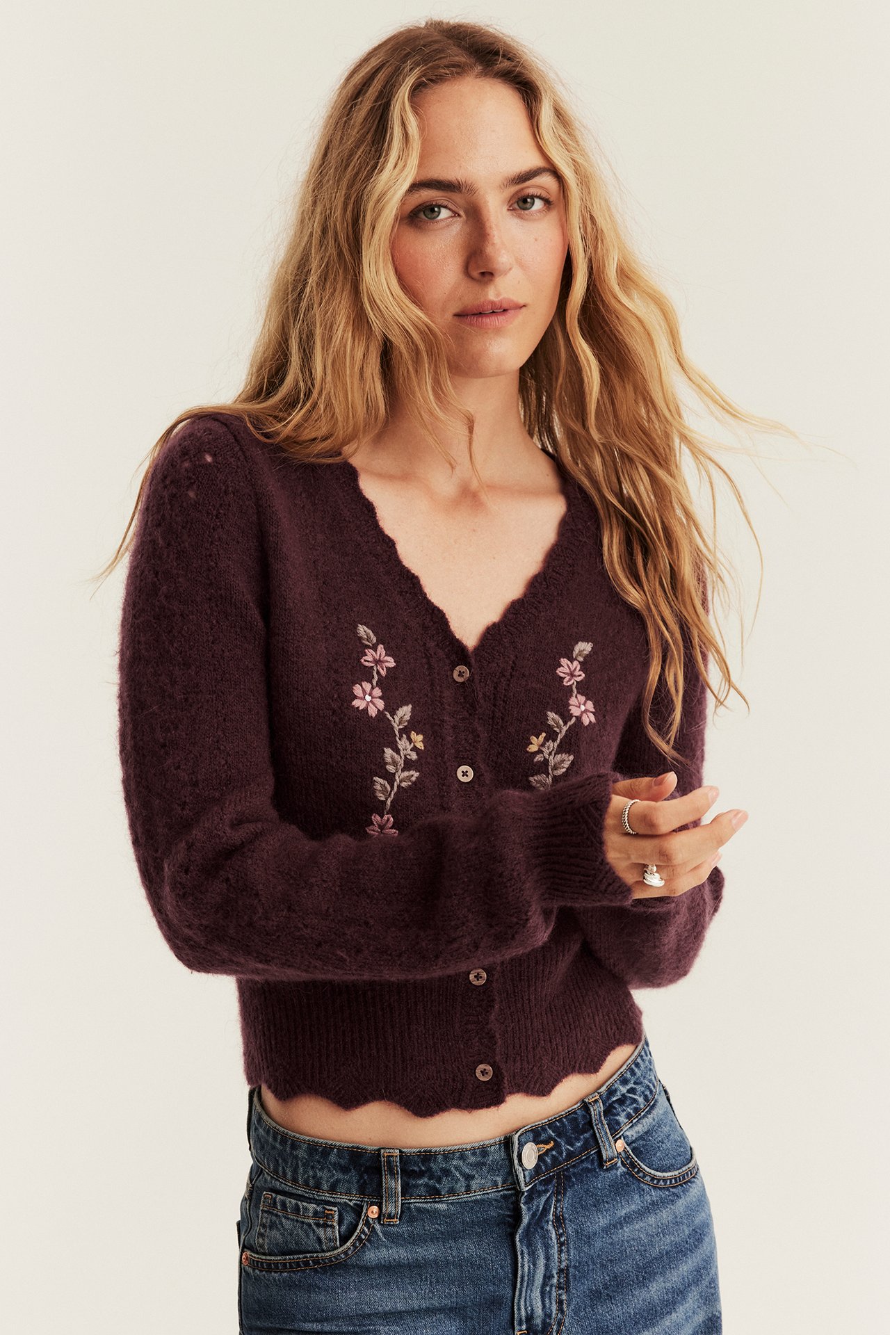 Vorderansicht einer Frau in dunkel pflaumenfarbener, flauschiger Strickjacke mit Blumenstickerei und blauen Jeans.