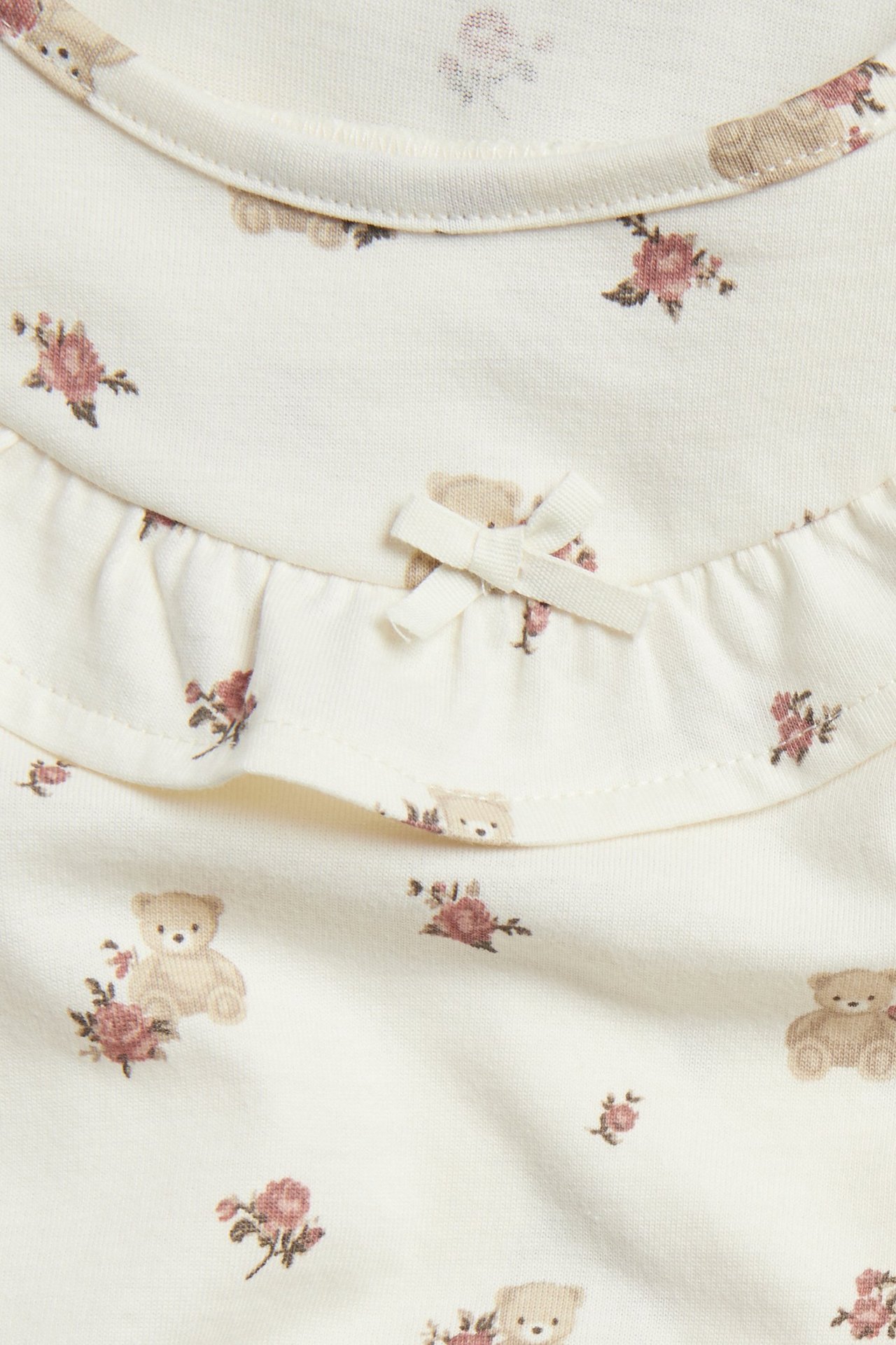 Nahaufnahme: cremefarbenes Baumwoll-Nachthemd mit Teddybär- und Rosenmuster, einer kleinen Schleife und Rüsche.