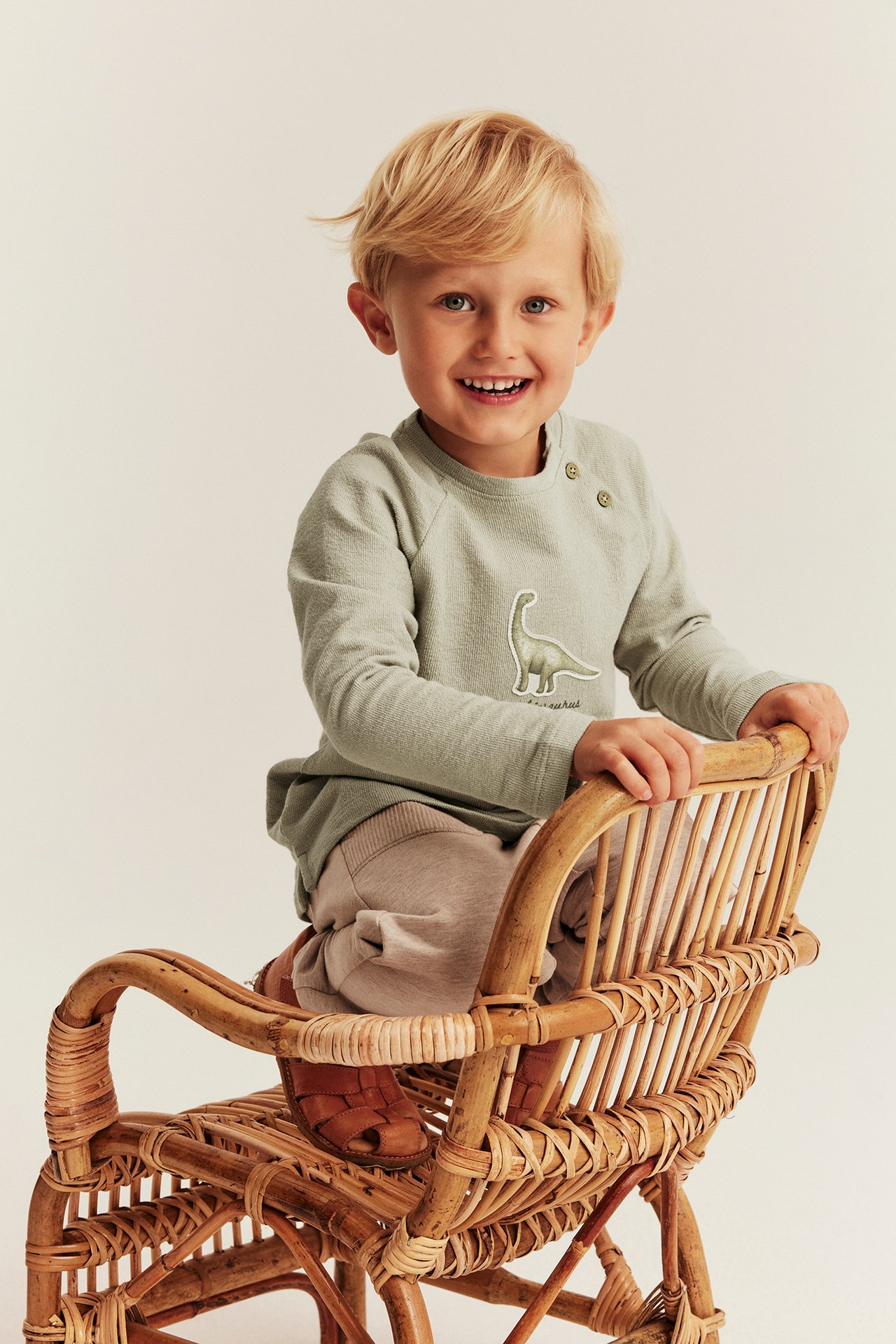 Front view of kid in light green structured top with dinosaur, beige trousers, brown sandals, on rattan chair.