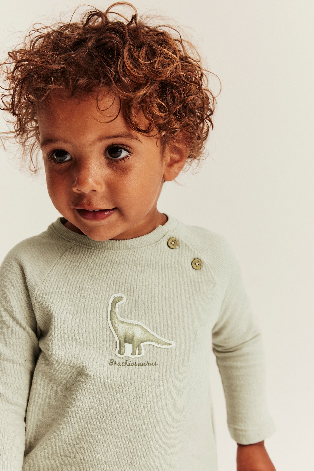 Front view of a kid wearing a light green long-sleeved structured cotton top with a dinosaur application and shoulder buttons.