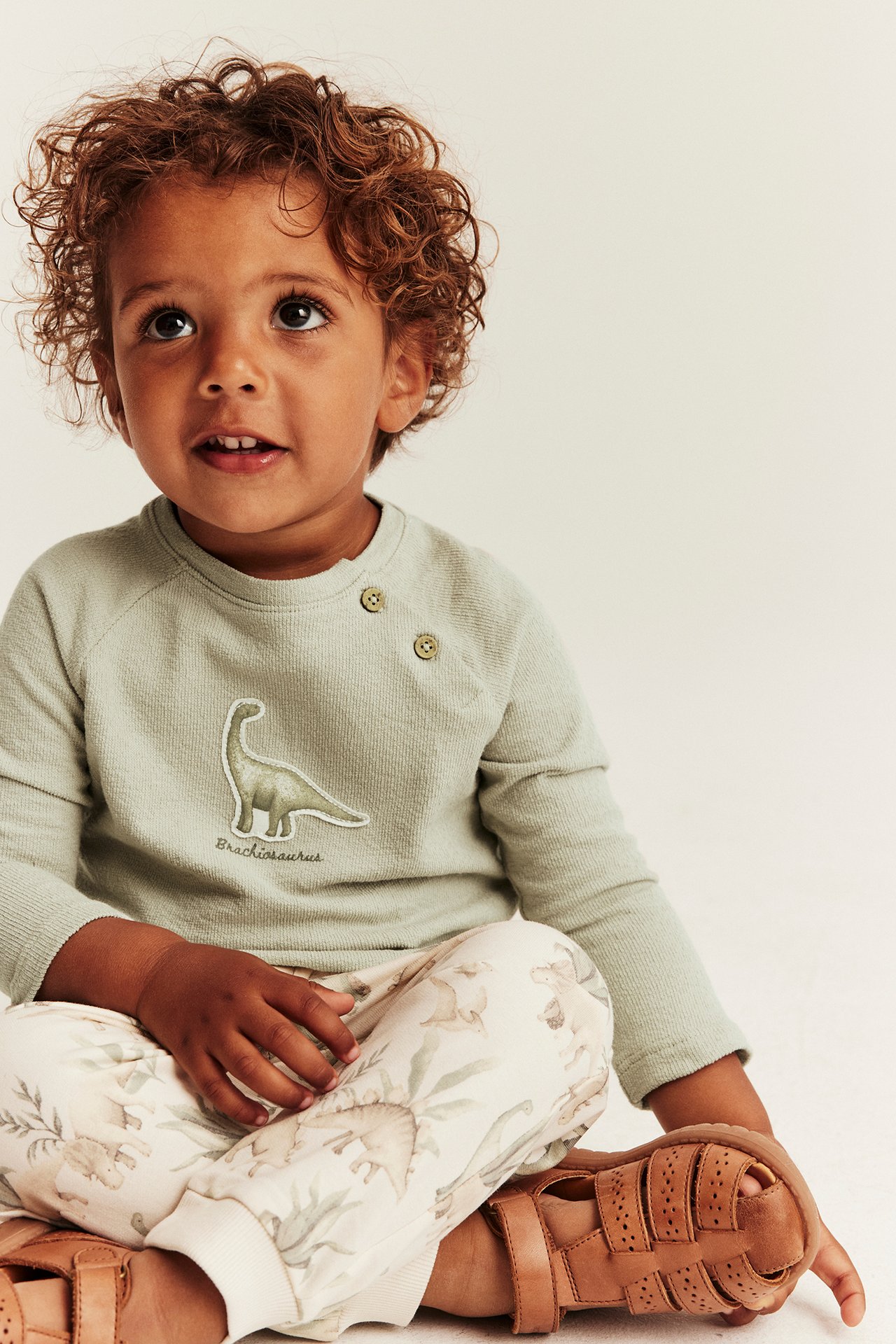 Front view: kid in light green long-sleeved top with dinosaur appliqué, patterned pants, and brown sandals.