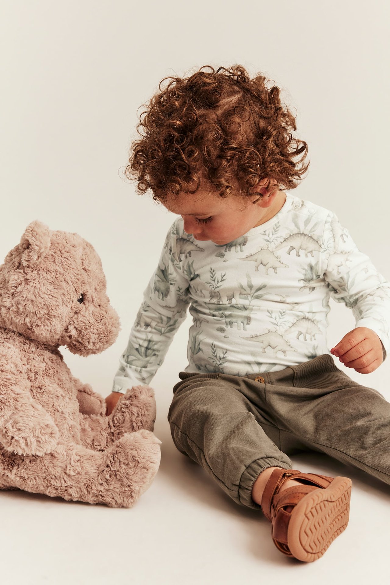 Ein Baby trägt ein weißes Langarmshirt mit Dinosaurier-Print, olivgrüne Jogginghose und braune Sandalen, nahe einem Teddybär.