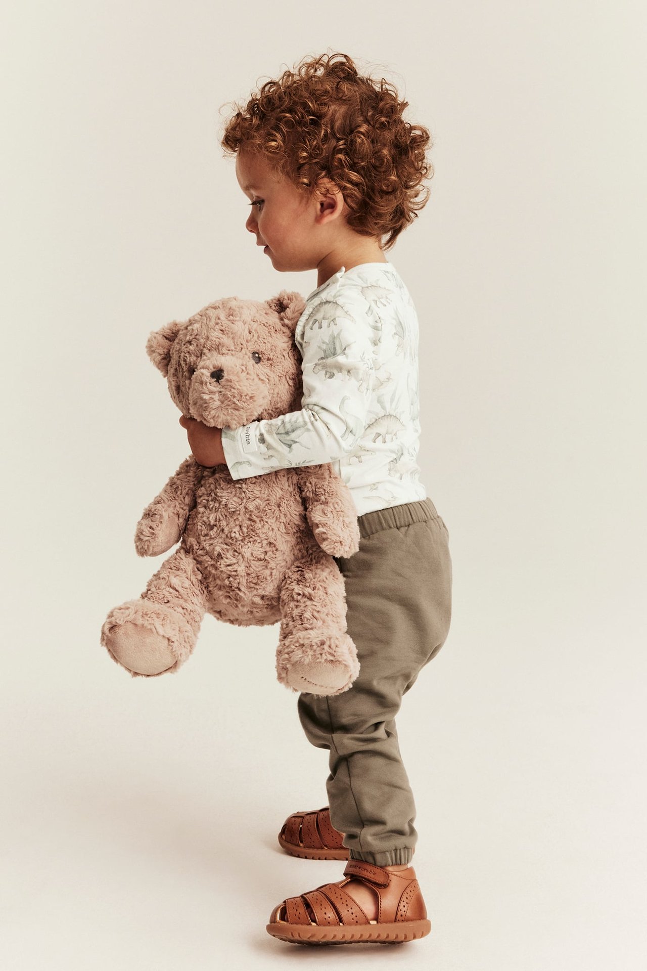 Kind hält hellbraunen Teddybär, weißes Dinosaurier-Oberteil, grüne Jogginghose, braune Ledersandalen, Seitenansicht.