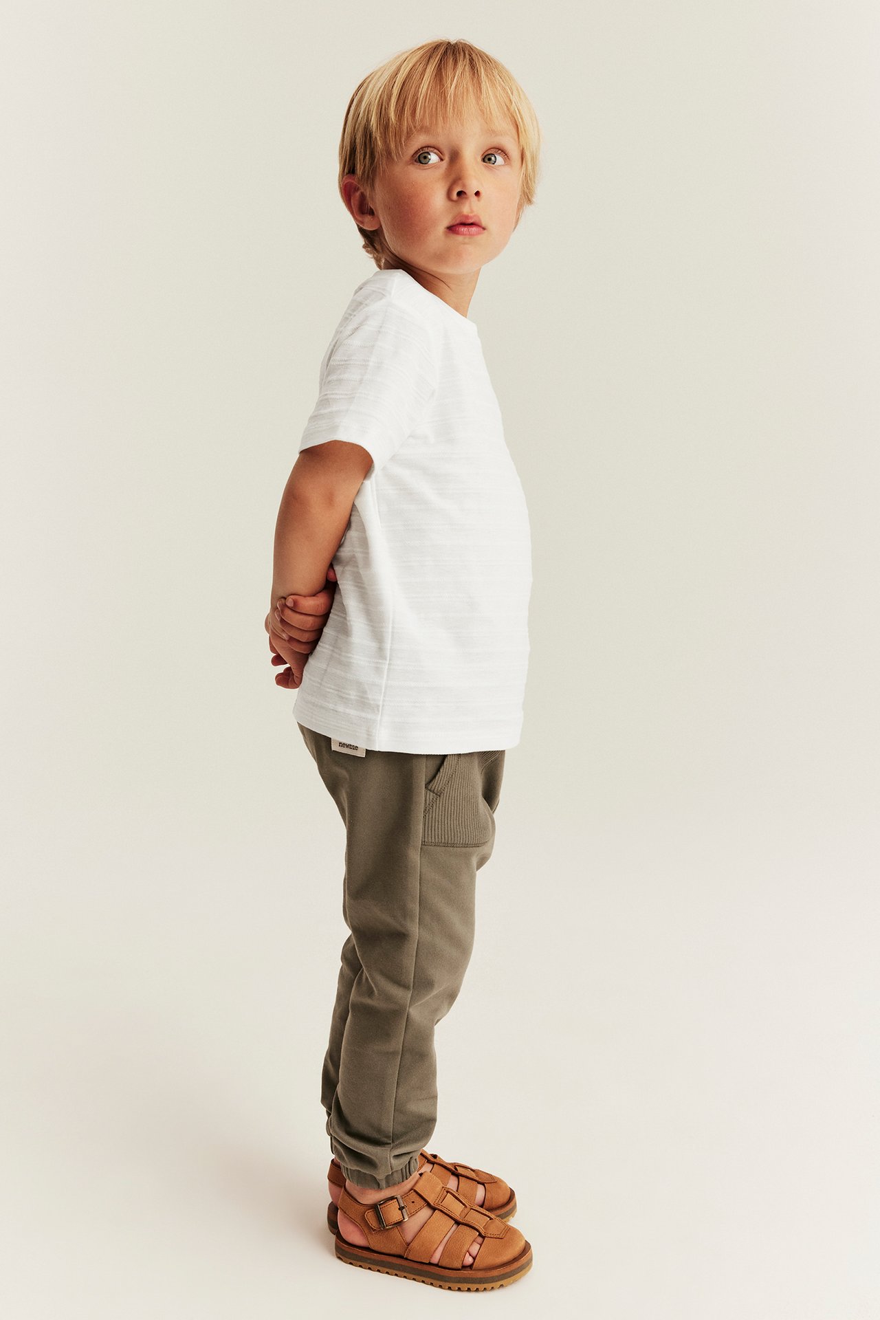 Side view of a kid wearing a white textured short-sleeve t-shirt, olive green sweatpants, and brown sandals.