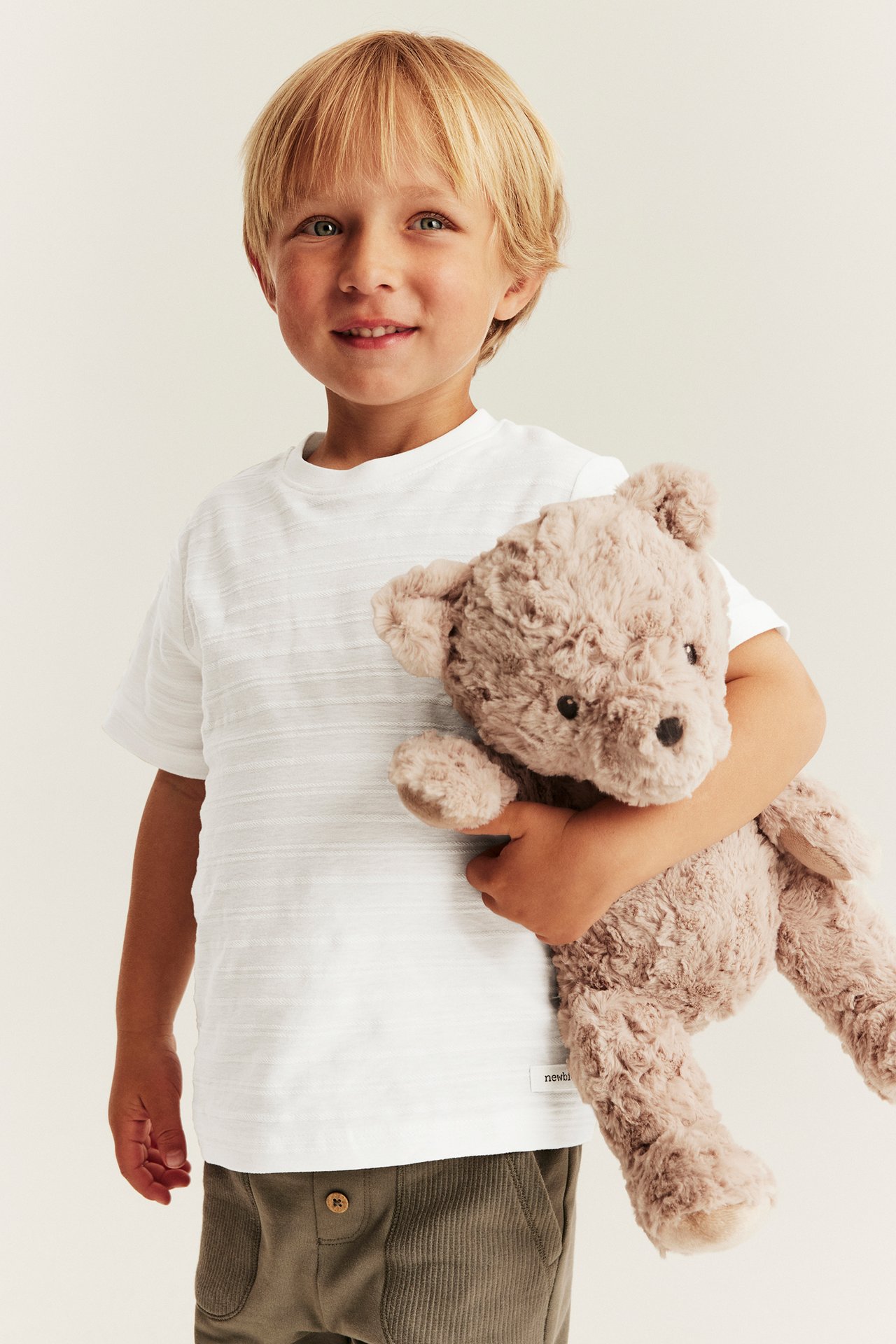 Front view of a boy wearing a white textured t-shirt and olive green ribbed trousers, holding a beige teddy bear.
