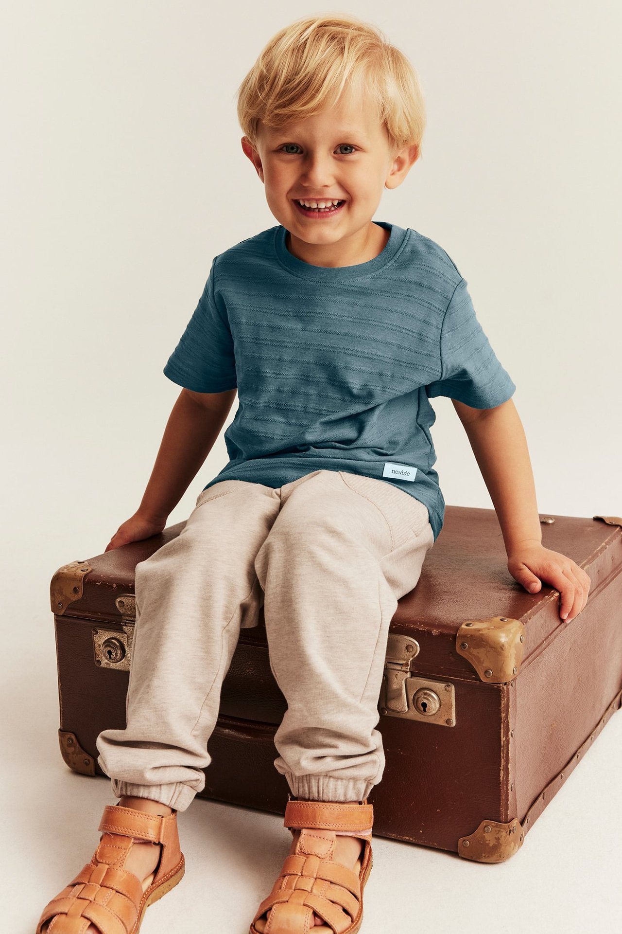 Vorderansicht eines Jungen in einem blau gestreiften T-Shirt, beiger Jogginghose und braunen Sandalen.