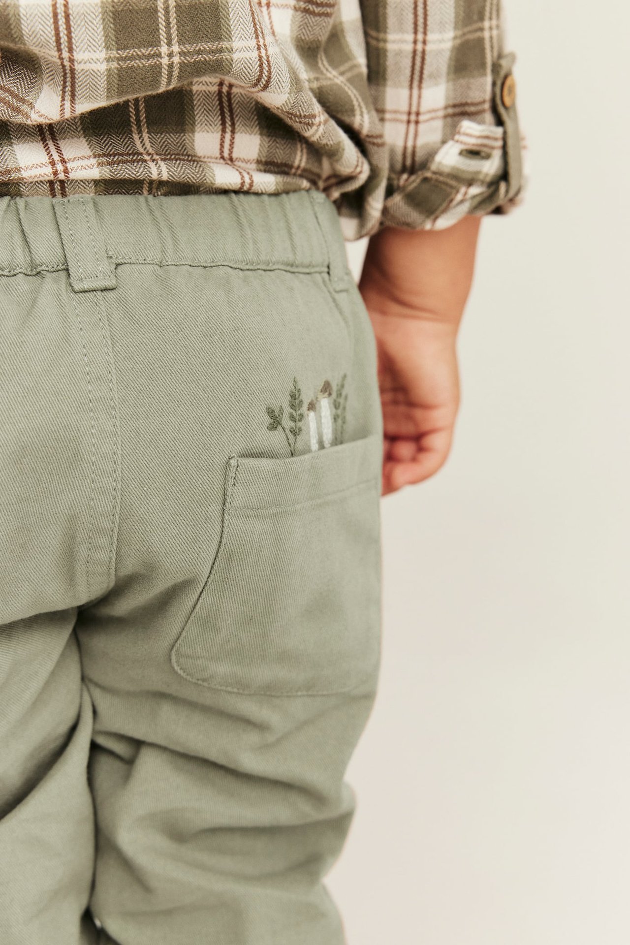 Nahaufnahme, Rückansicht eines Kindes in olivgrüner Hose mit bestickter Tasche und einem grün karierten Hemd.