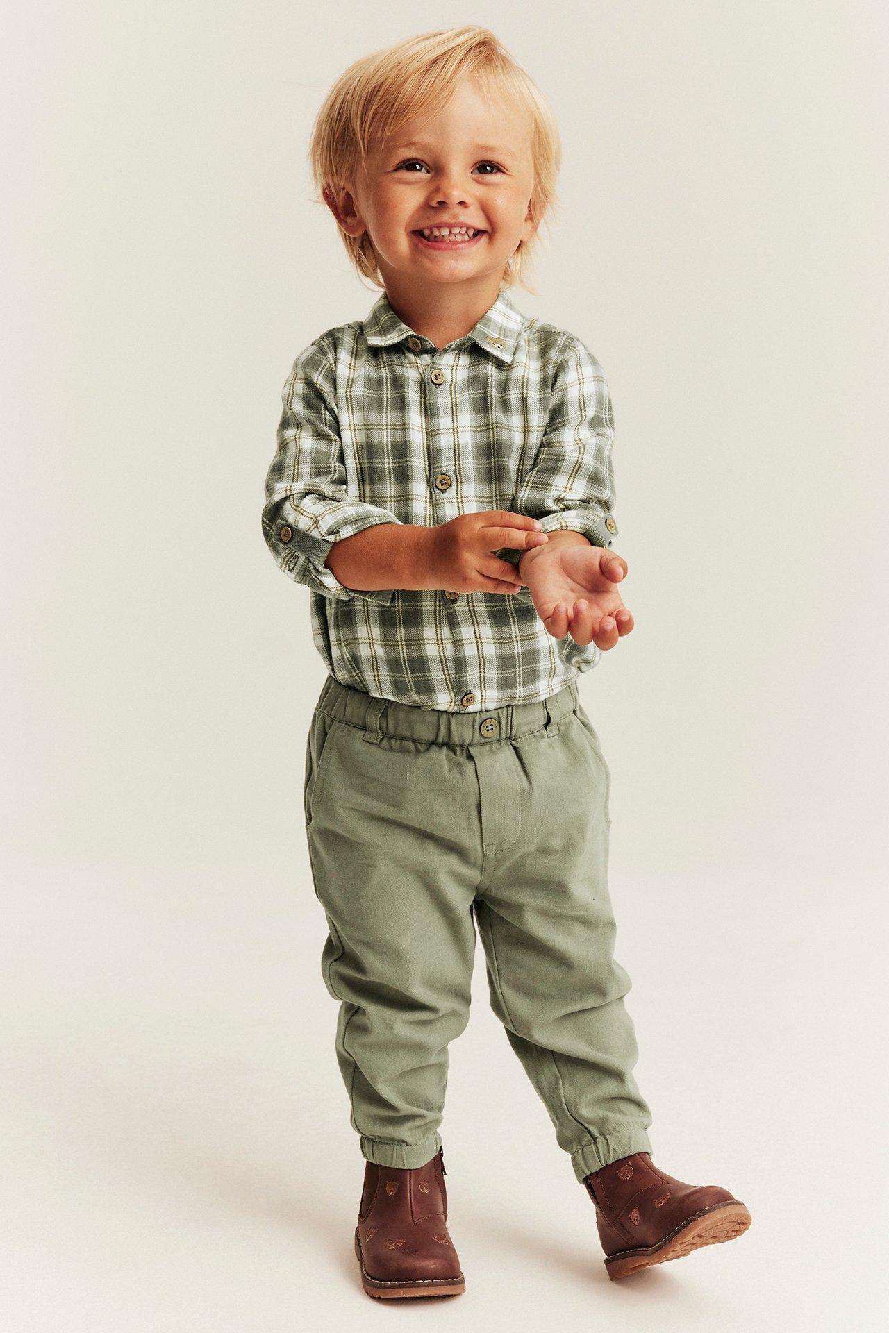 Front view of a kid wearing a green plaid shirt, sage green joggers, and brown boots.