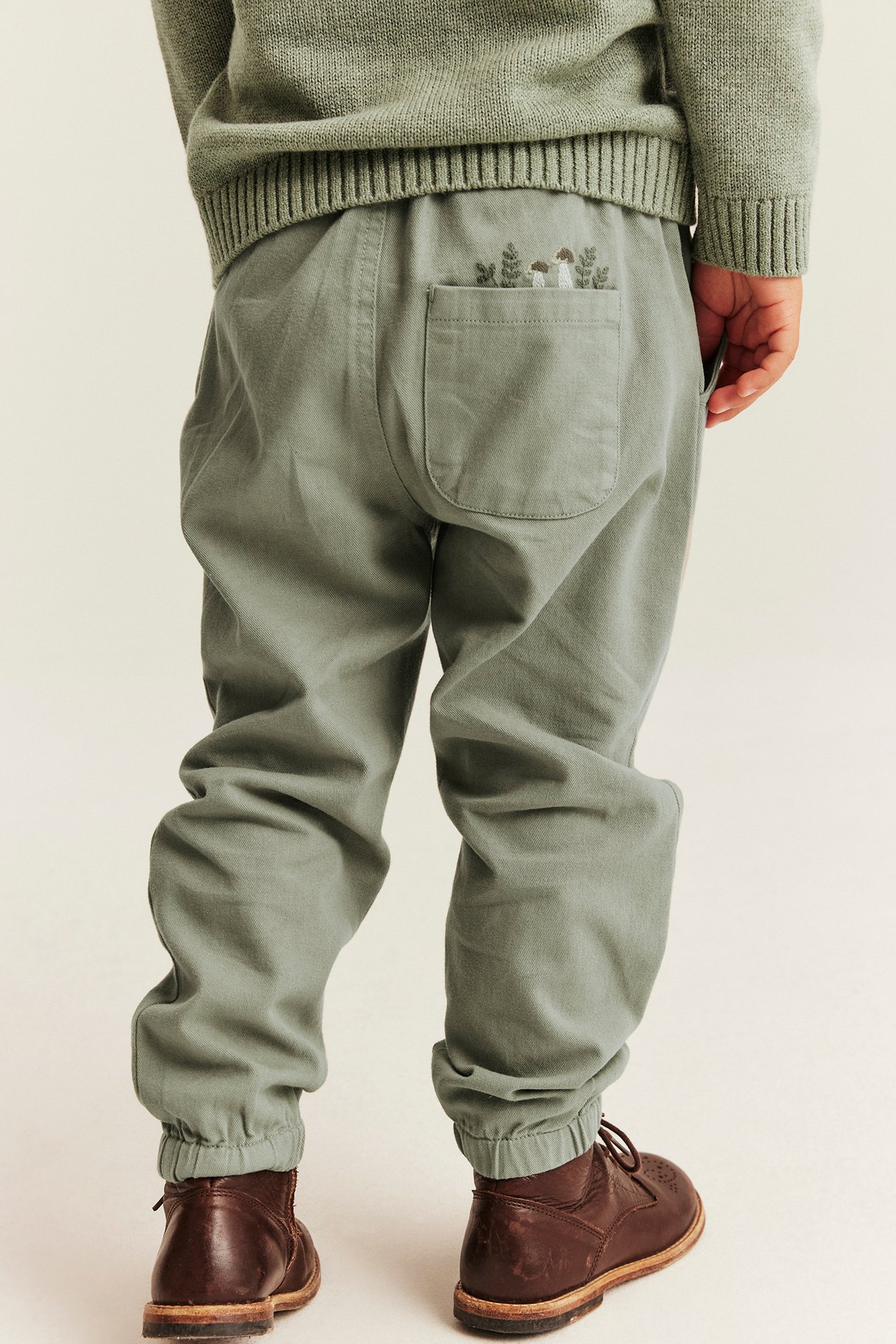 Back view of kid wearing green pants with mushroom embroidery, matching sweater and brown boots.