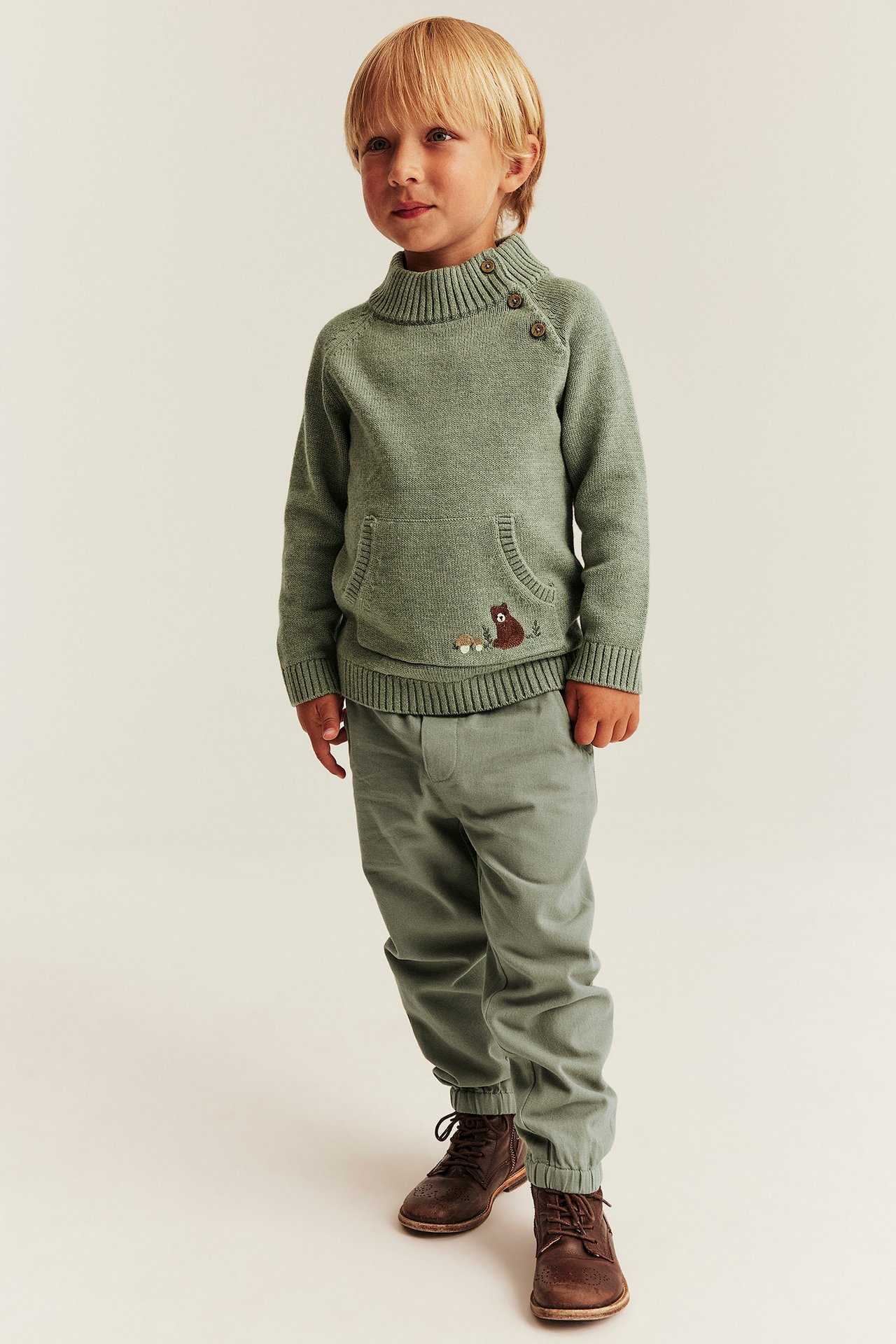 Vorderansicht: Kind in salbeigrüner Strickpullover mit Knopfkragen, grüner Hose und braunen Stiefeln.