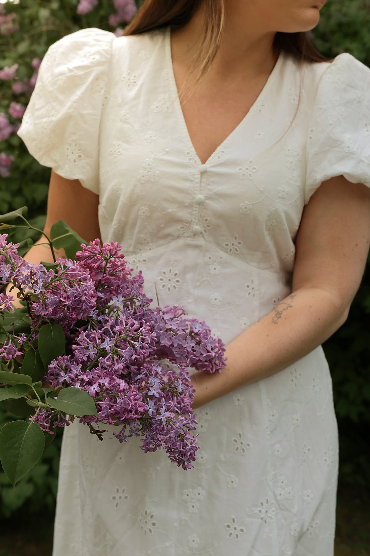Close-up front view: woman in offwhite embroidered cotton dress with short puff sleeves, holding purple lilacs.