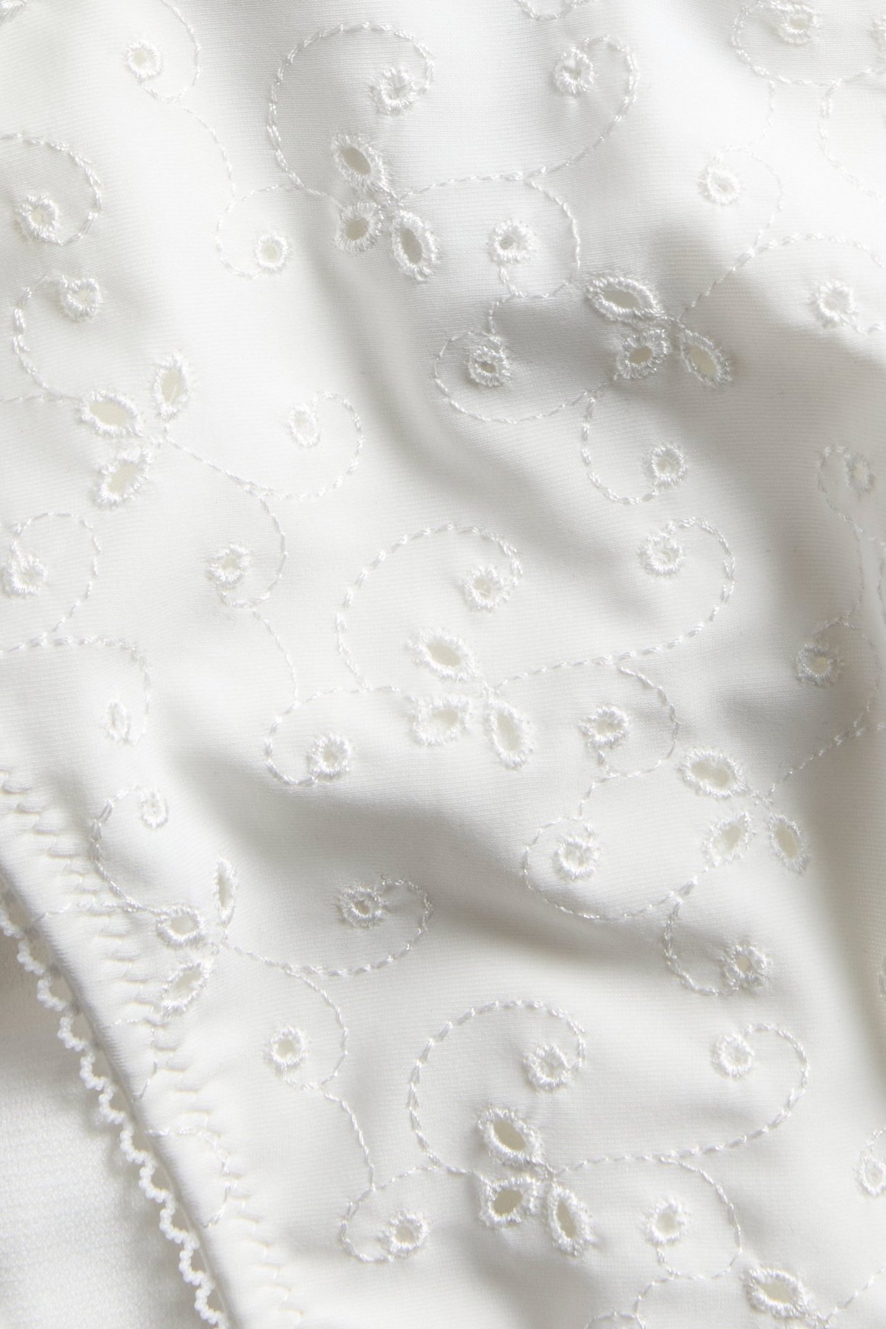 Close-up of off-white bikini bottom fabric with delicate all-over white embroidery and a decorative scalloped edge.