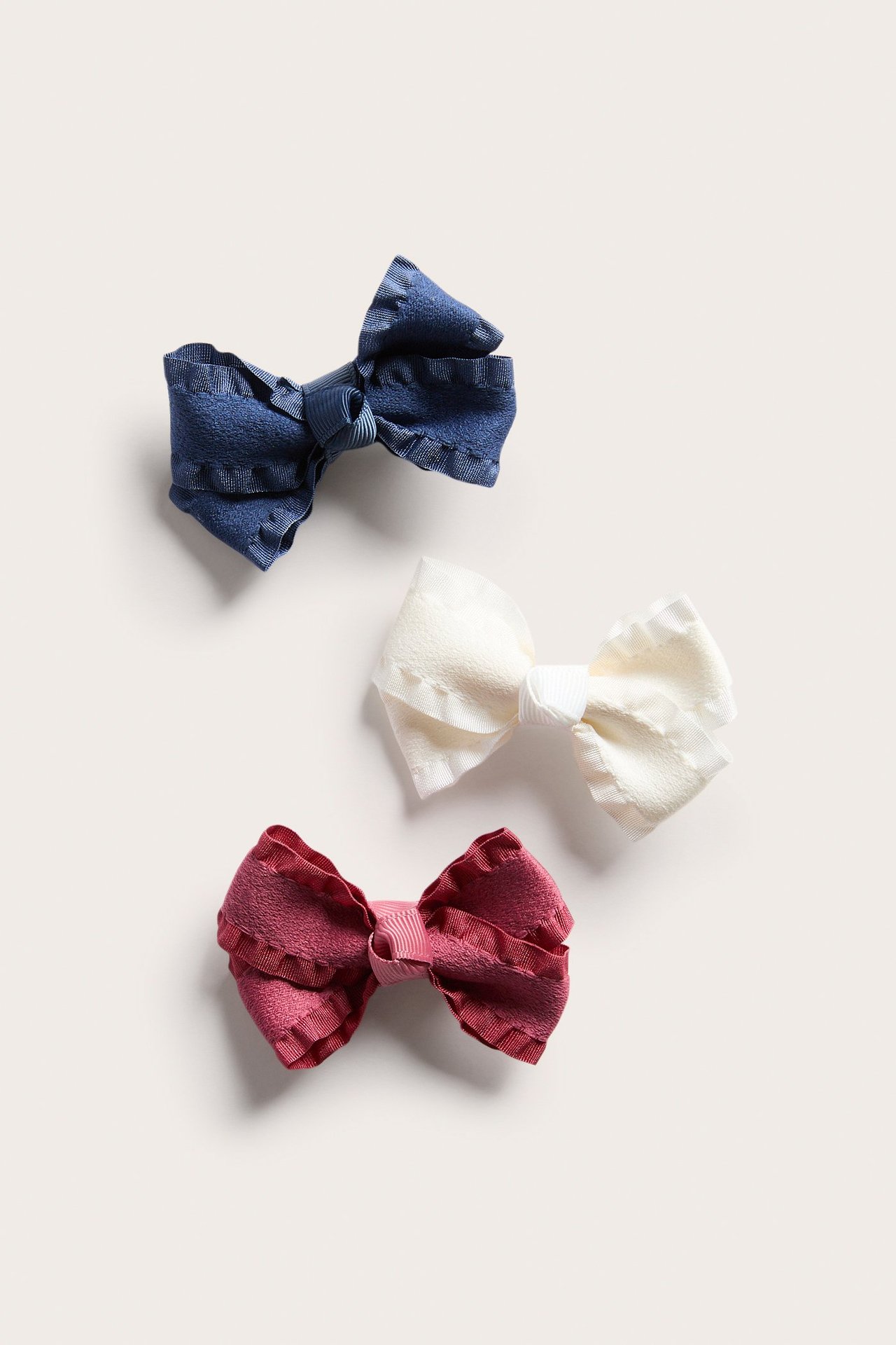 Top view of three textile bow hair clips in dark blue, off-white, and berry red with ruffled edges.