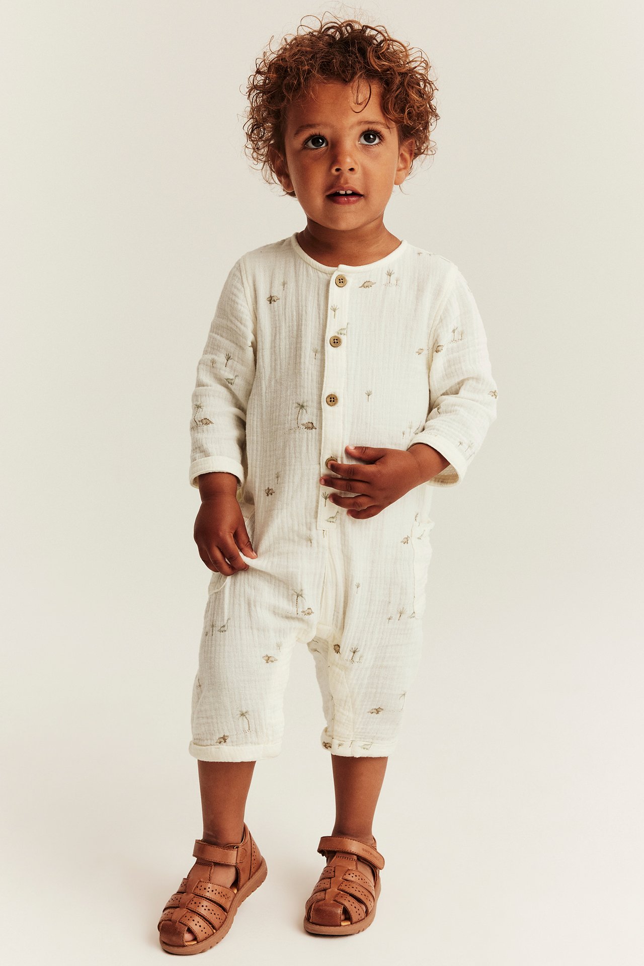 Vorderansicht eines Babys in cremefarbenem Musselin-Overall mit Palmen- und Dinosaurier-Print und braunen Sandalen.
