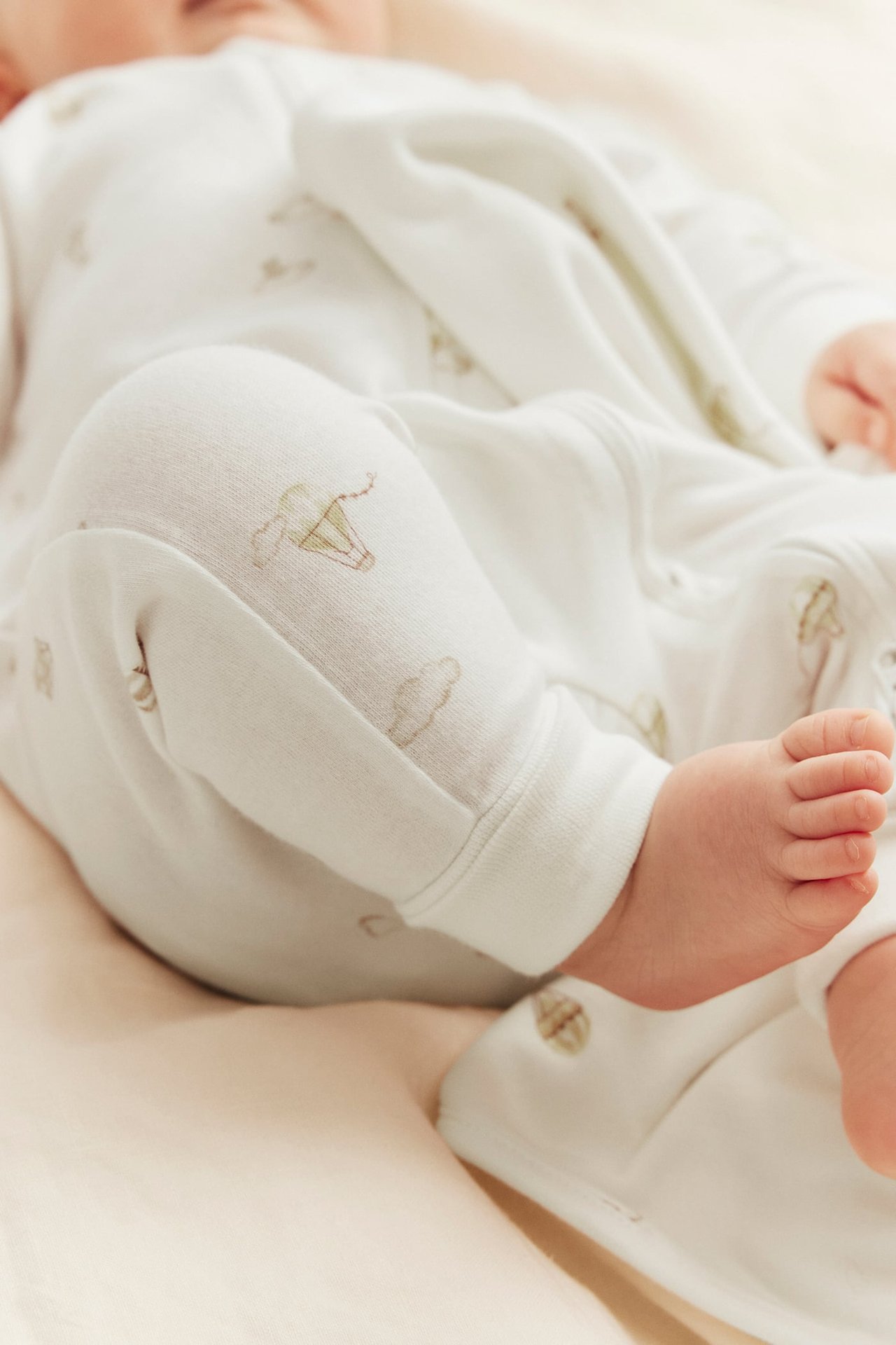 Nahaufnahme: Baby in cremeweißem Baumwoll-Strampler mit Heißluftballons, Frontreißverschluss und umklappbaren Bündchen.