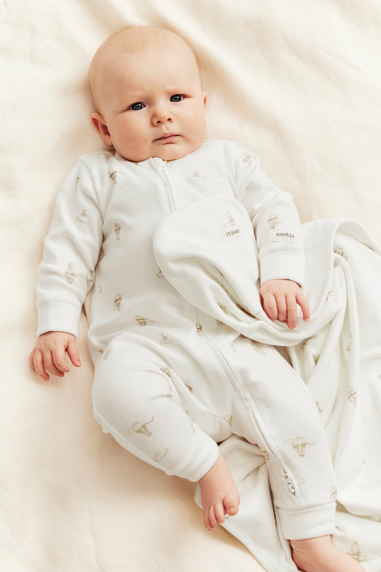 Aufsicht: Baby in cremeweißem Strampler mit Heißluftballonmuster, Frontreißverschluss. Passende Decke.
