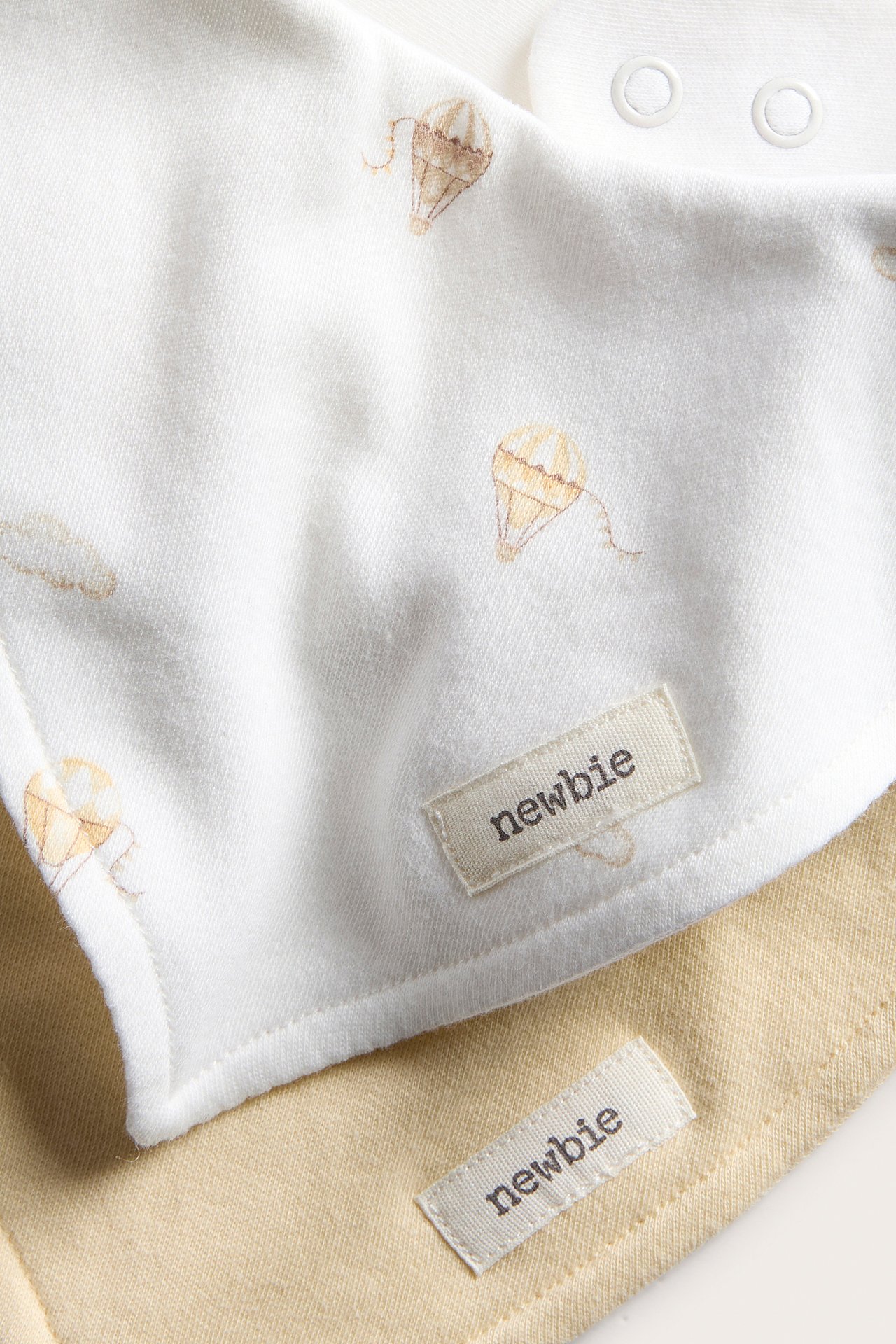 Nahaufnahme von zwei Baby-Baumwolllätzchen: eines cremeweiß mit Heißluftballons und Druckknöpfen, ein hellbeiges.