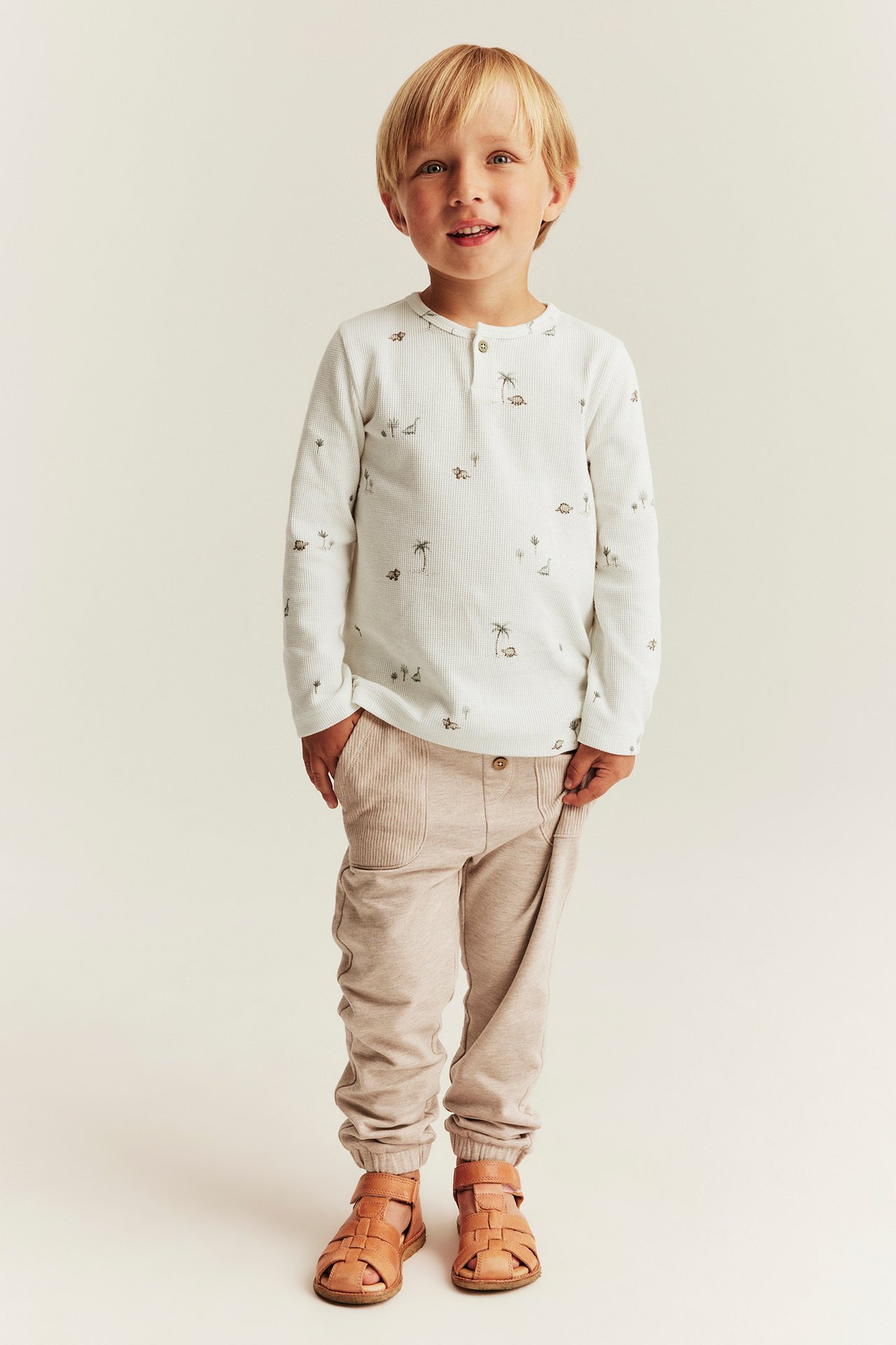 Vorderansicht: Kind in offweißem Waffel-Langarmshirt mit Knopfleiste, Dino-Muster, beiger Hose und braunen Sandalen.