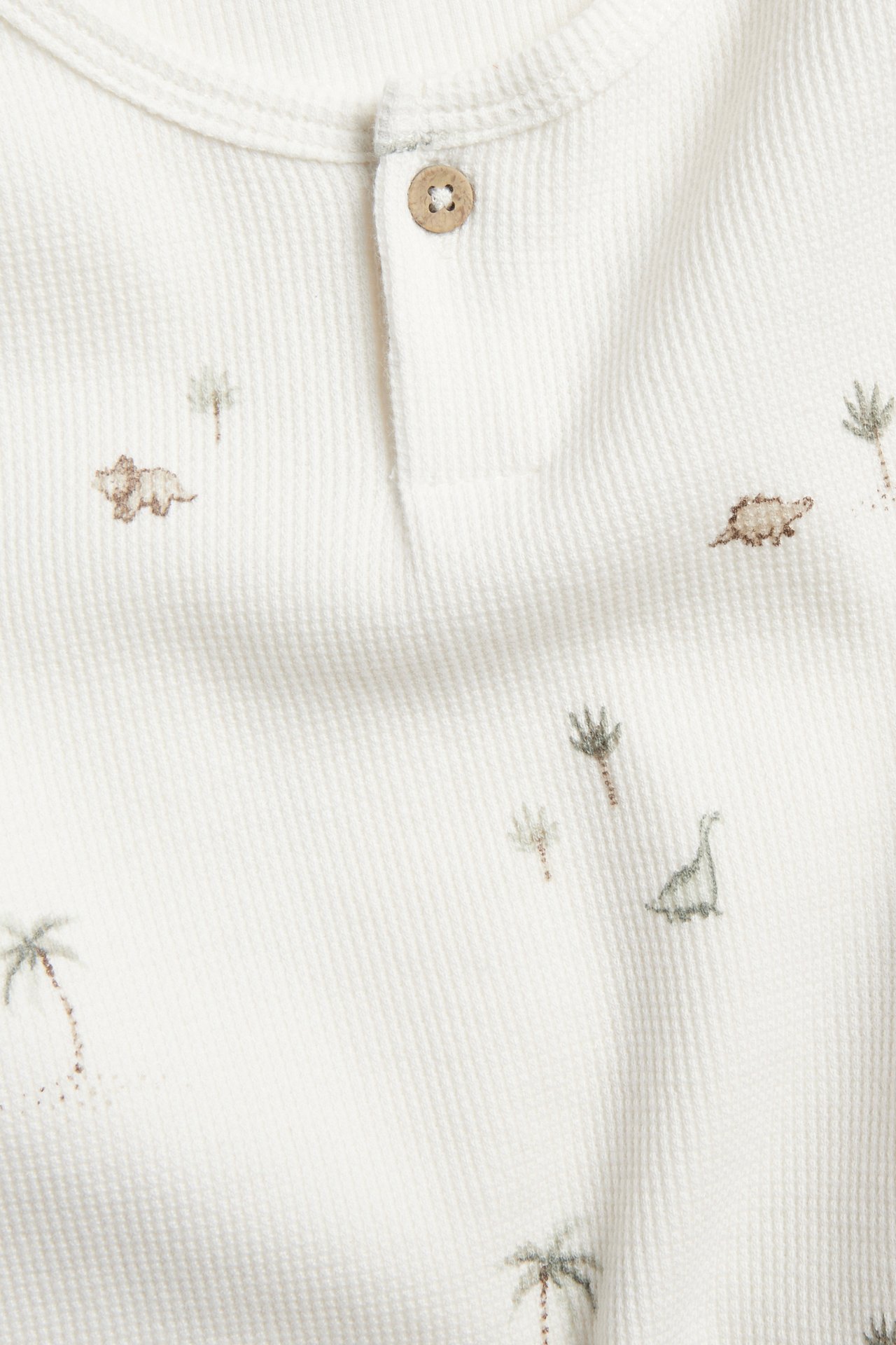 Close-up: Off-white waffle long-sleeved t-shirt with button placket, dinosaur and palm tree pattern.