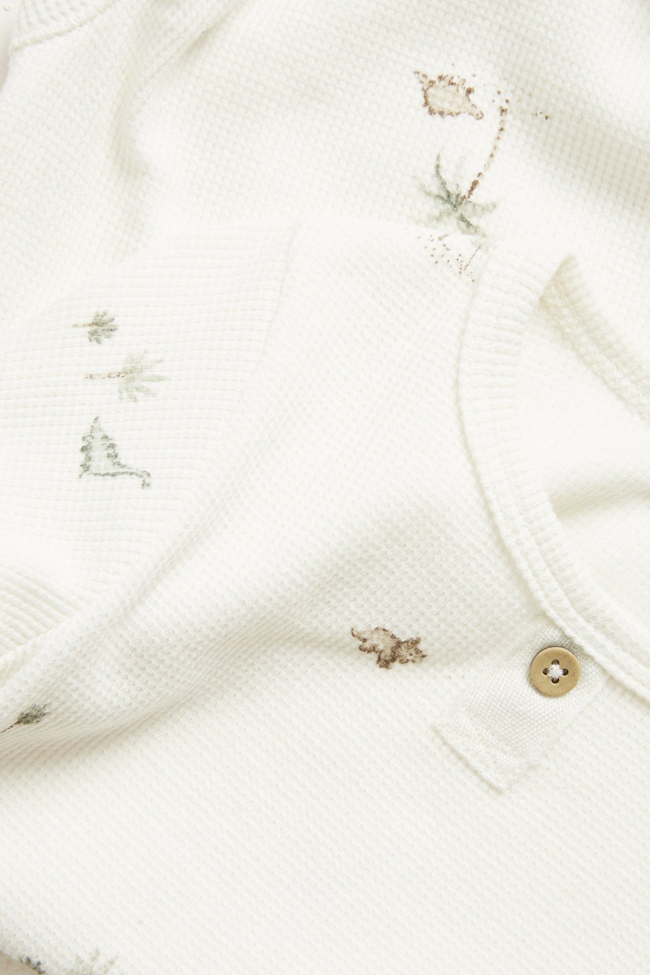Nahaufnahme cremeweißen Waffelstrick-Baby-Langarmbodys mit gestickten Dinosauriern, Palmen und Zierknopf.