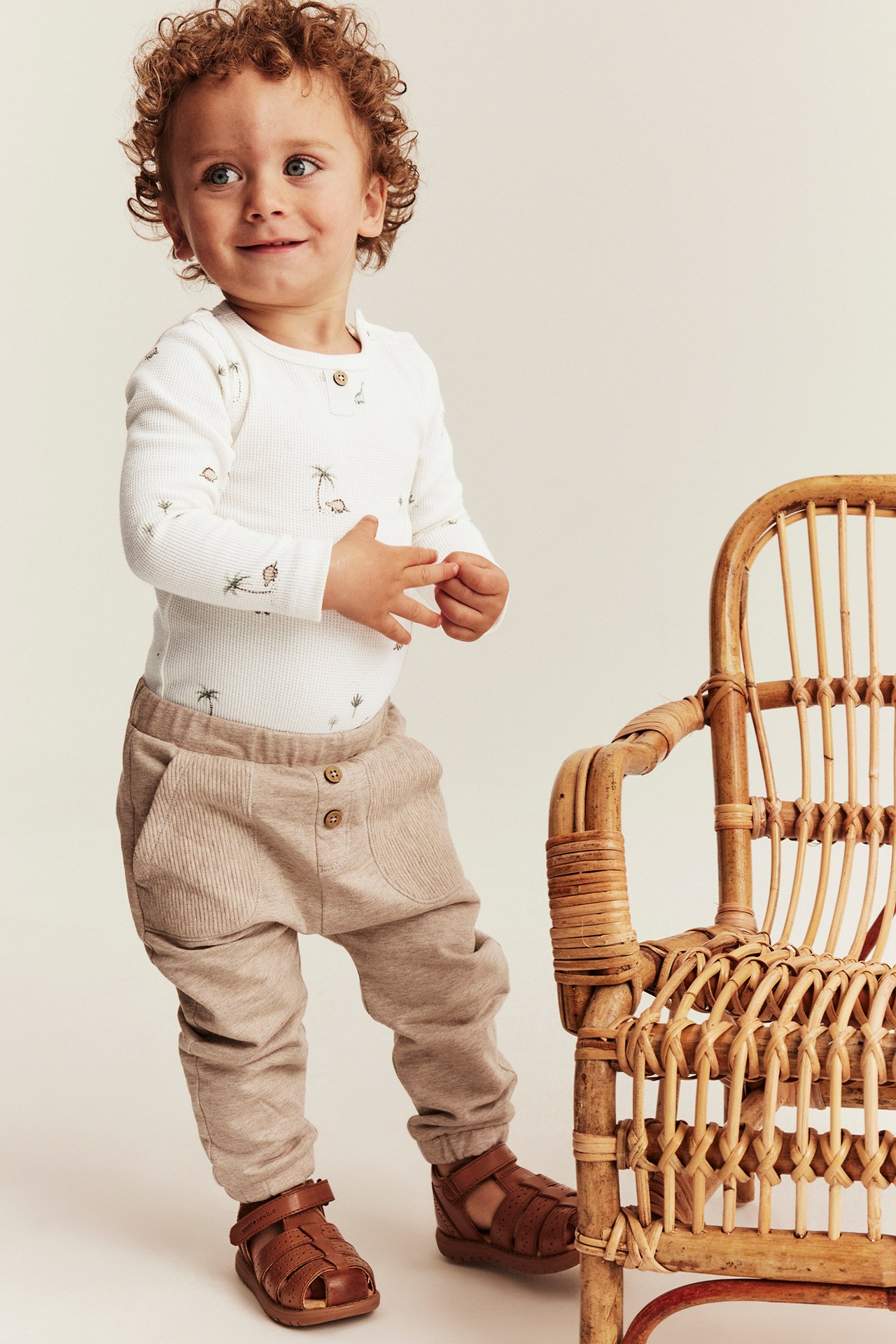 Vorderansicht, Baby in einem cremeweißen Waffel-Langarmbody mit Dinosaurier-Print, beiger Ripphose und braunen Ledersandalen.
