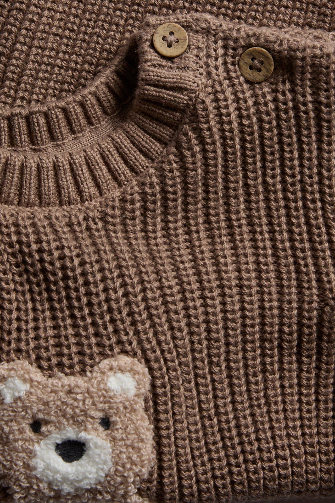 Close-up of a brown knitted baby sweater with wooden buttons and a teddy bear appliqué.
