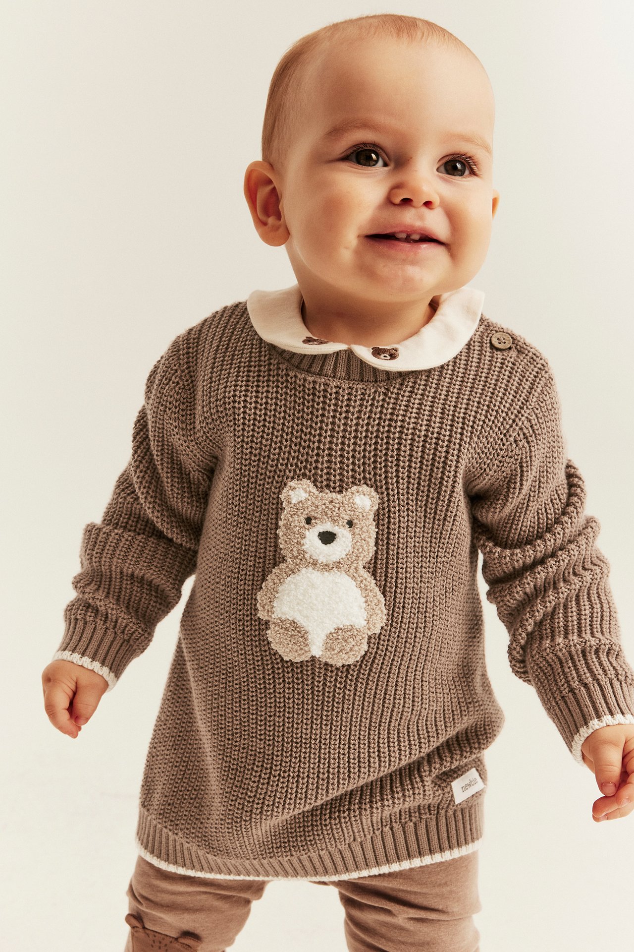 Front view of a smiling baby wearing a brown knitted teddy bear sweater, white collar, and brown leggings.