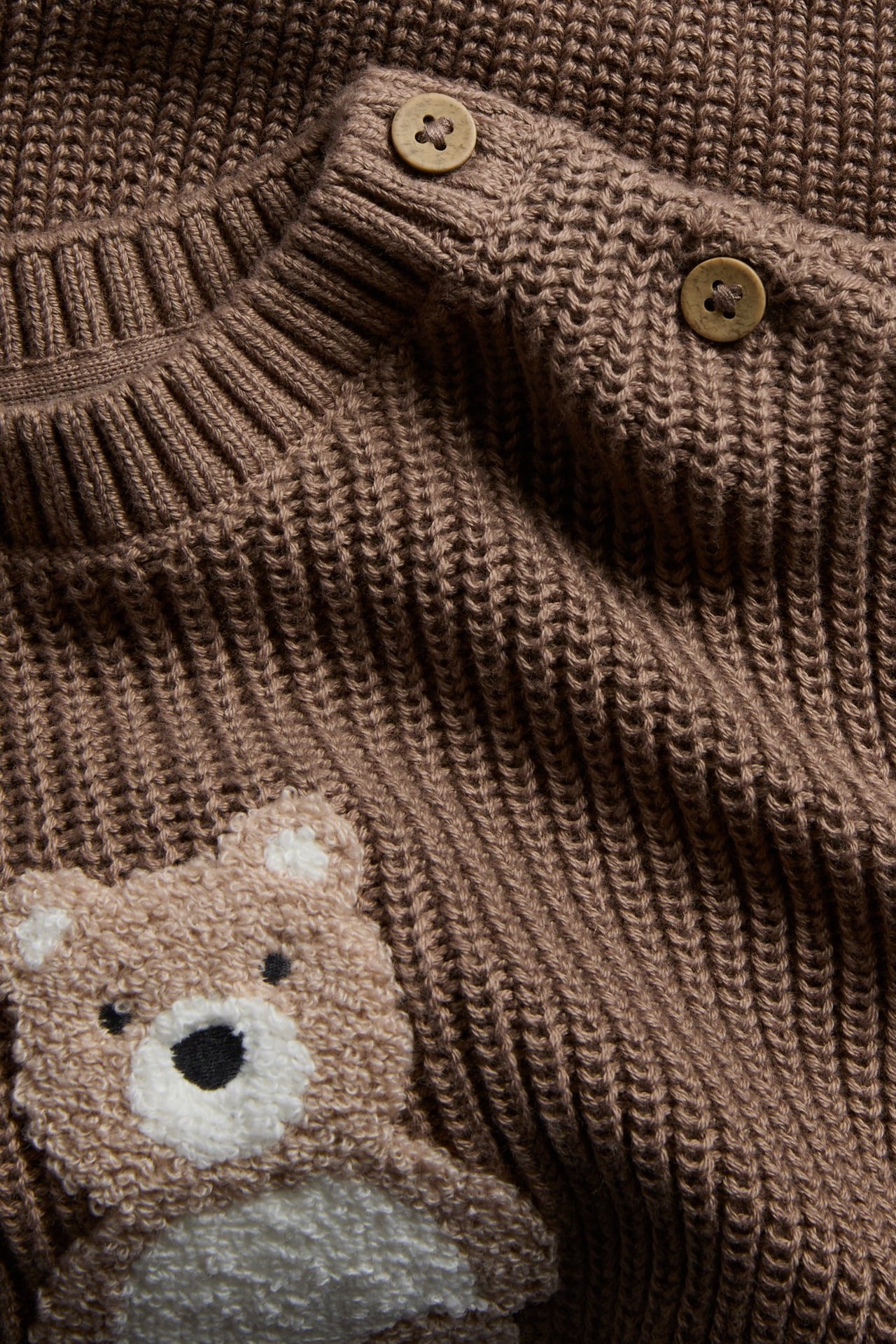 Nahaufnahme eines braunen gestrickten Baby-Pullovers mit Teddybär-Applikation und zwei Knöpfen.