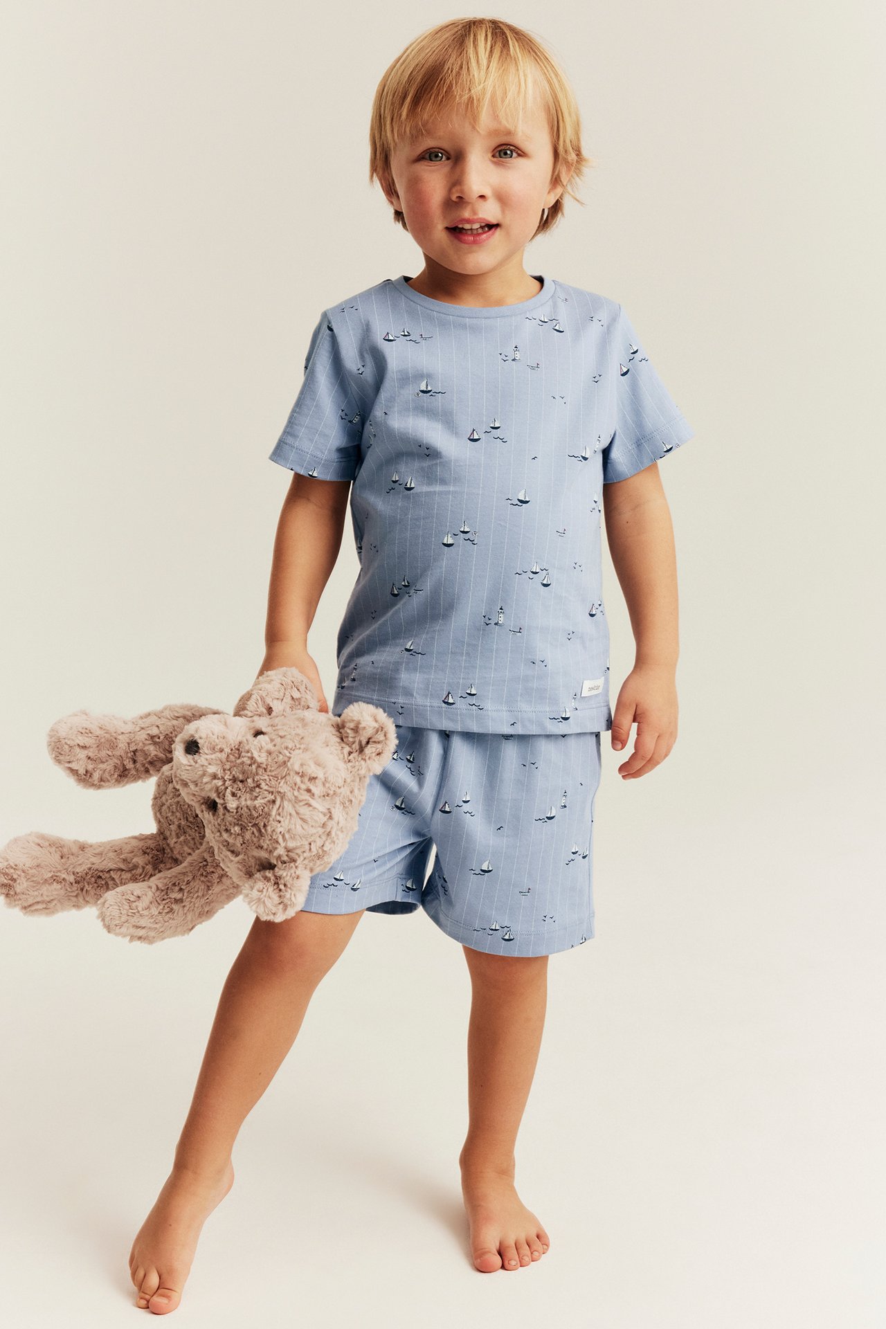 Kid in light blue cotton pyjama set with short sleeves, boat pattern. Holds brown teddy bear.