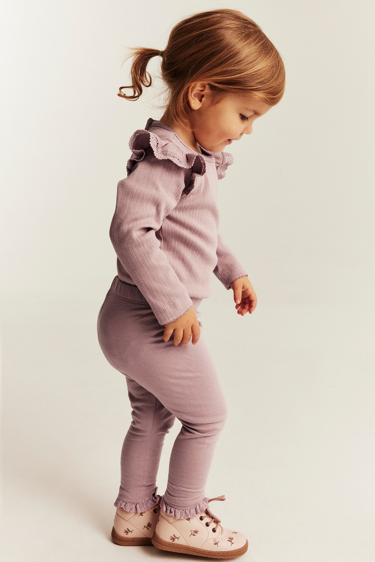 Kind in staubigem lila Rippentop mit Rüschen, passenden Leggings und rosa Blumenschuhen.