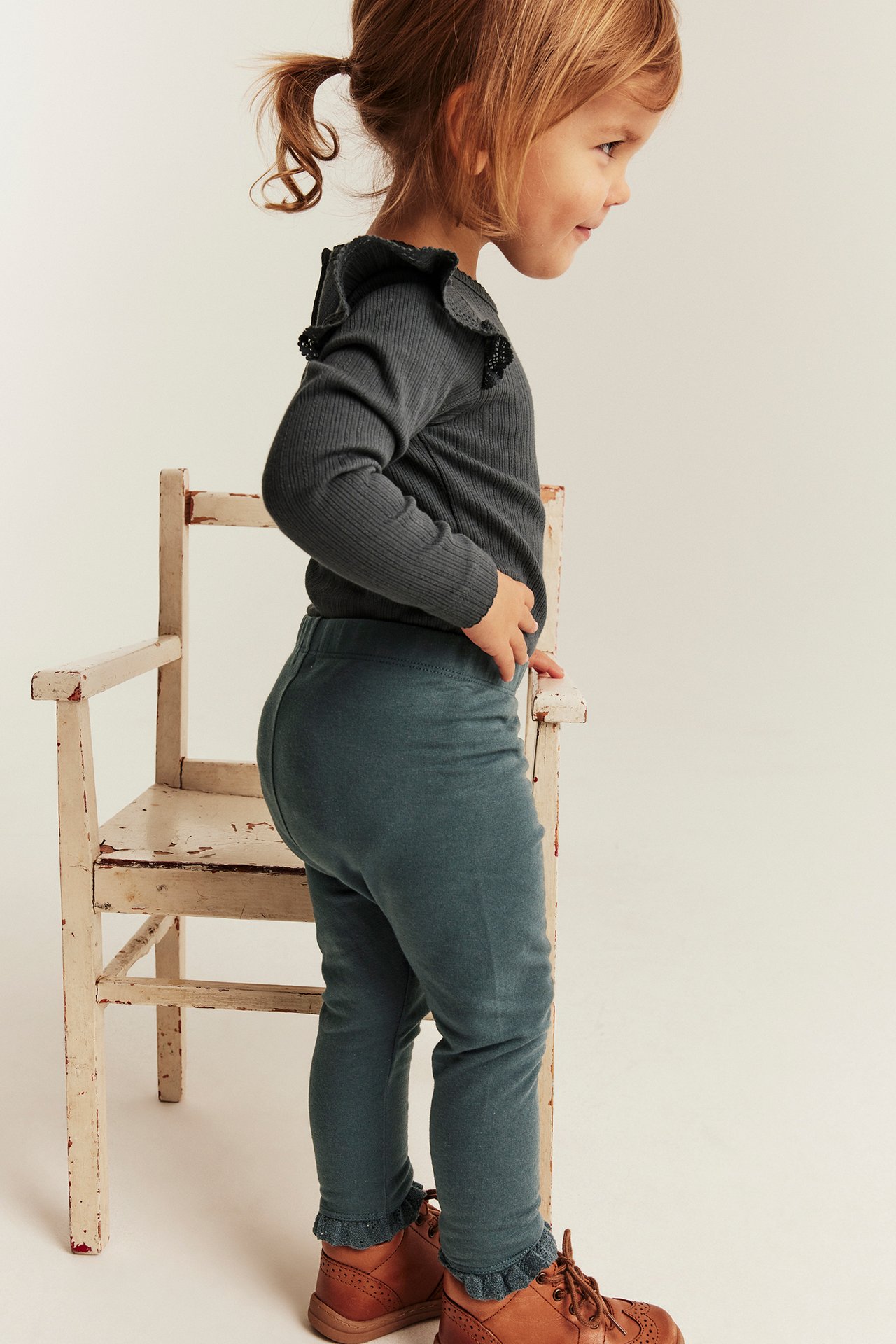 Seitenansicht Kind in teal Leggings, einem dunkelgrauen Ripp-Top mit Rüschen und braunen Stiefeln.