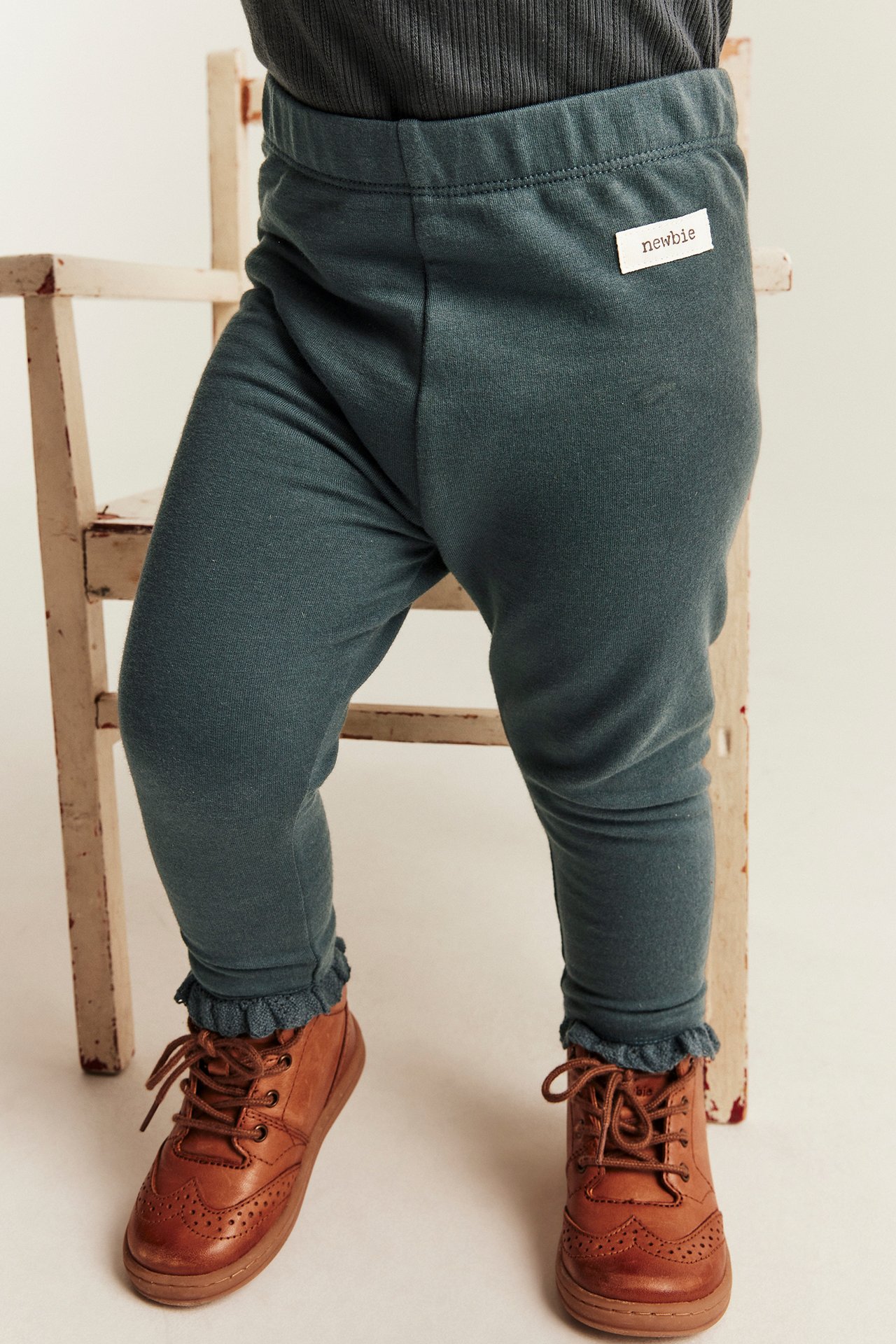 Baby trägt dunkelblaue Jersey-Leggings mit Rüschensaum, dunkelgraues Top und braune Schnürstiefel.