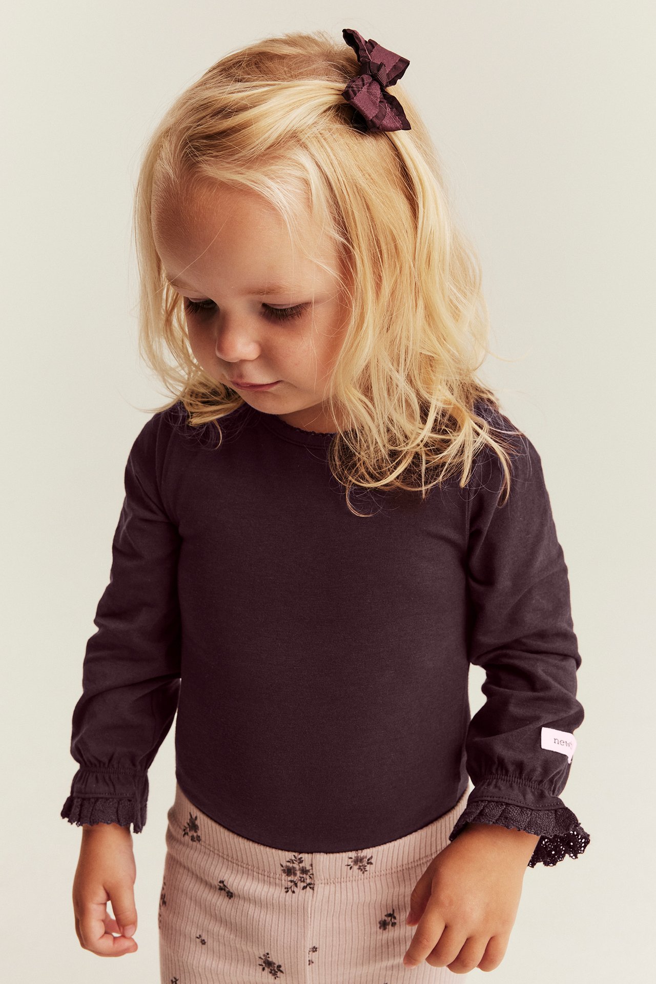 Vorderansicht eines Kindes in einem dunkelvioletten Langarmshirt mit Rüschen und Spitze sowie bedruckter beiger Hose.