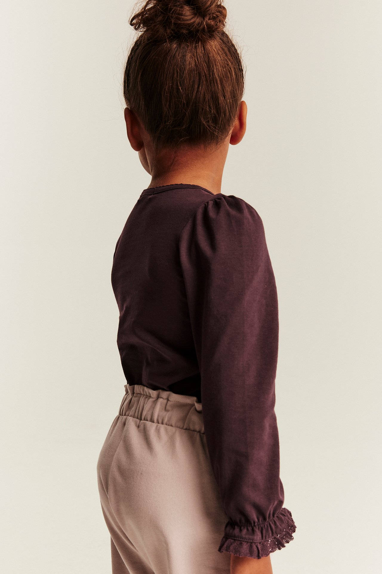 Rückenansicht eines Kindes in dunkelviolettem Langarmshirt mit Puffärmeln und beiger Hose.
